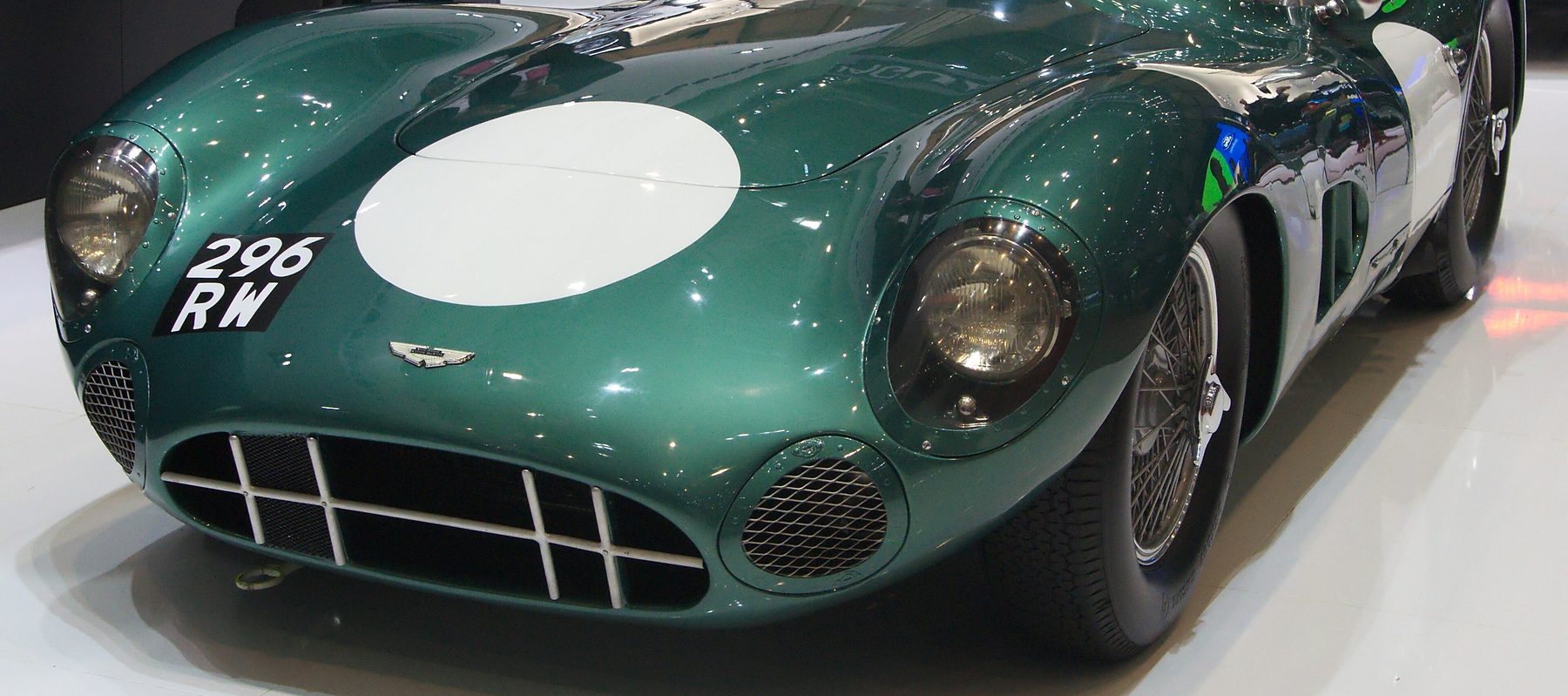 The nose of a gleaming dark green open sports-racing car, a white roundel on its bonnet and the registration 296 RW, on a pale show-stand floor.