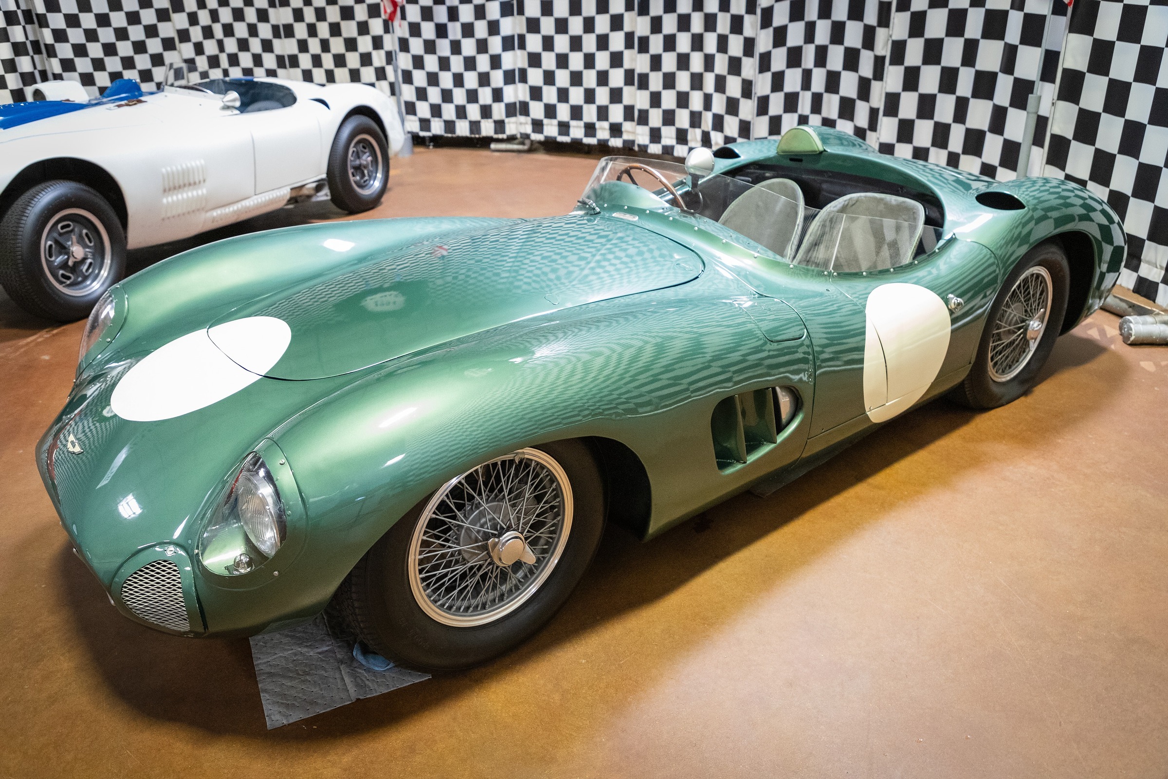 A long, curved, green open sports-racing car with white roundels on its bonnet and flank stands on a polished floor before chequered black and white drapes; part of a white sports car shows at the left.