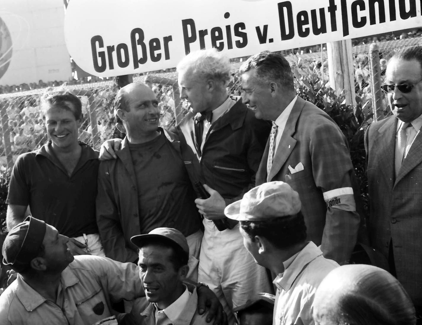 Three drivers stand under a race banner among officials and mechanics: a young man smiling at the left, an older man in the centre, and a fair-haired man in a dark jacket beside him.