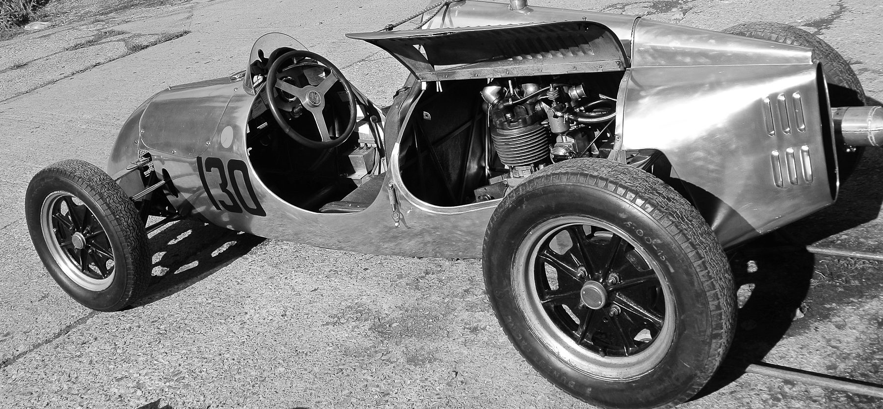 A small polished open single-seater numbered 130, its engine cover raised over a single-cylinder engine behind the seat, stands on a concrete paddock in bright sun.