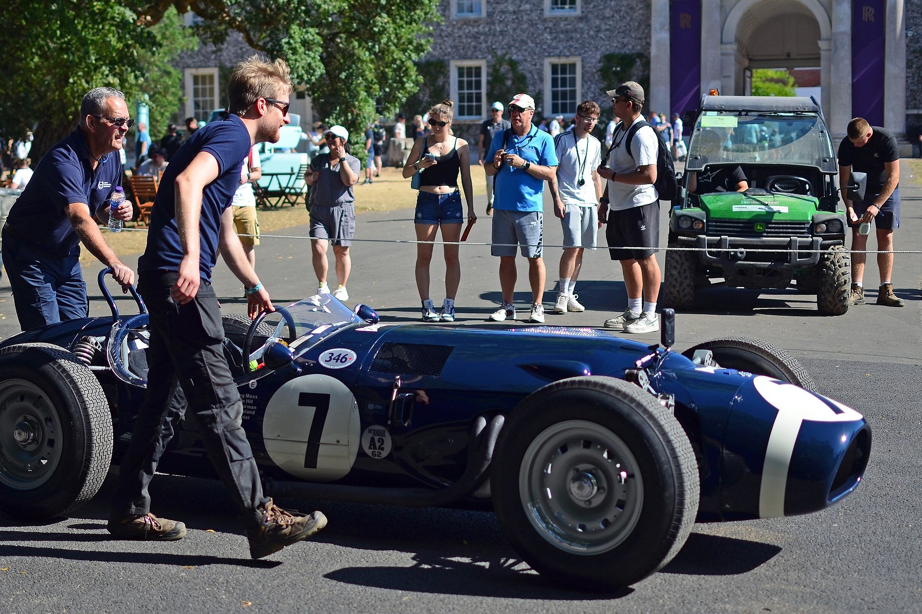 Two men in dark shirts push a small dark blue front-engined racing car, numbered 7 and with a white band round its nose, across a paddock; onlookers stand behind a rope and a utility vehicle is parked at the right.