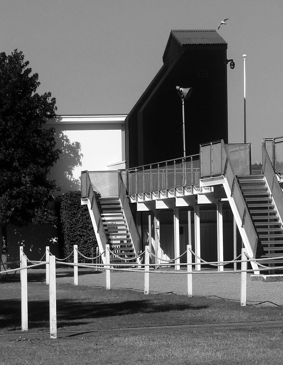 Two open flights of stairs climb to an empty grandstand beside a tall, dark, windowless tower; a rope fence on white posts and a strip of grass in front, a tree at the left and a gull in the sky.