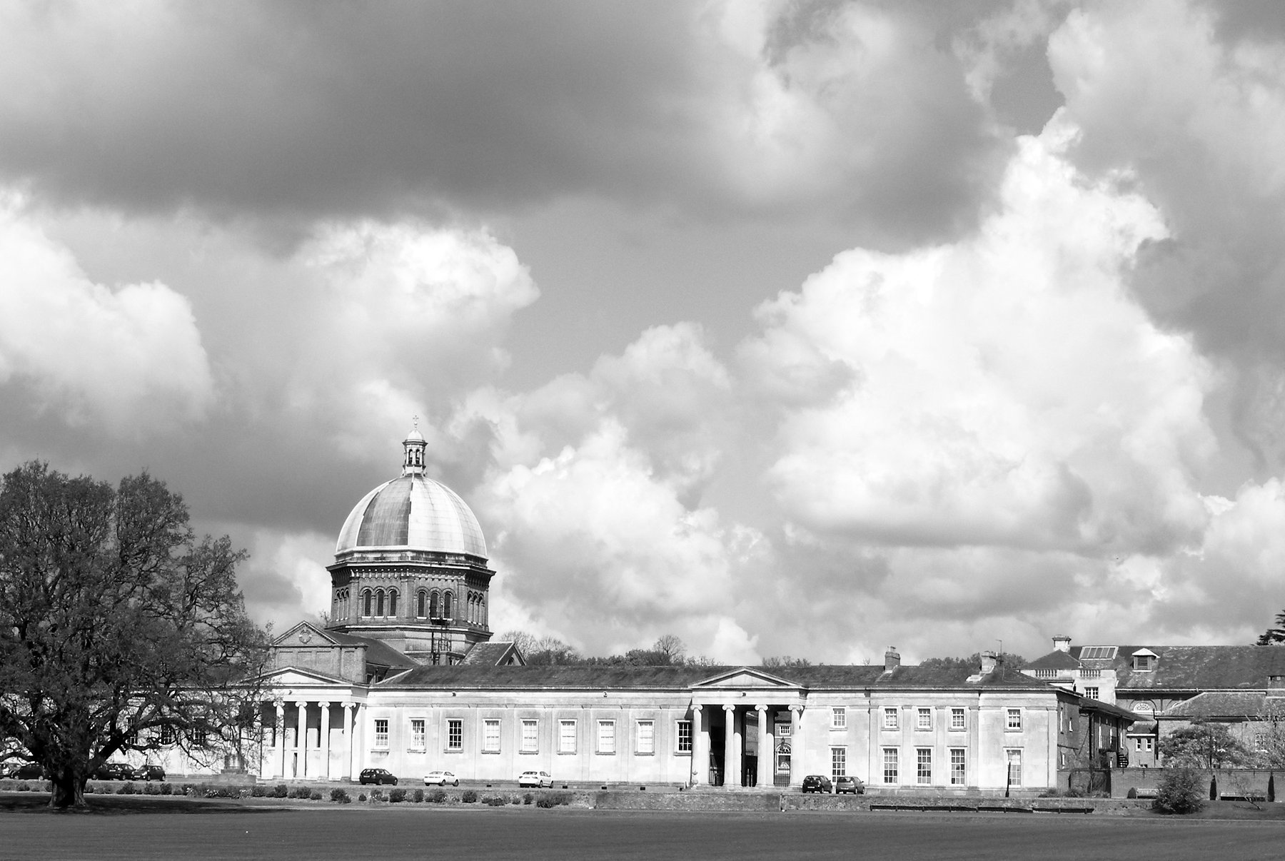 A long colonnaded building with a domed chapel stands beyond wide playing fields under a towering clouded sky, a large oak at the left.