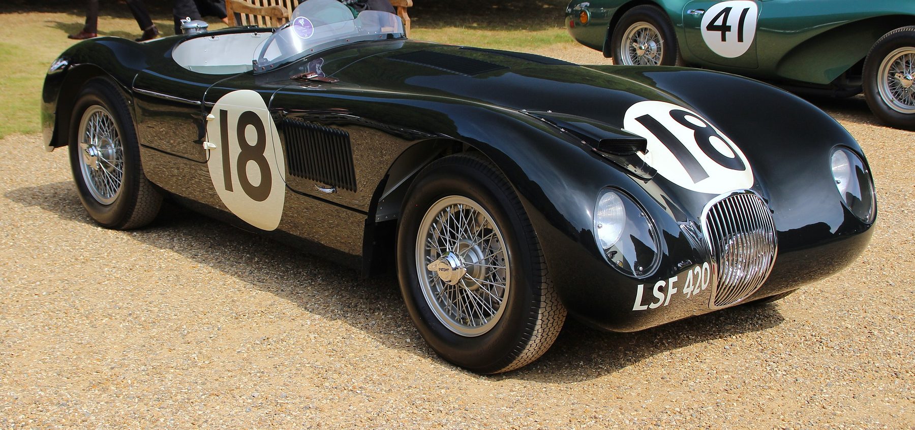 A low, rounded dark green sports-racing car numbered 18, registration LSF 420, stands on gravel; a second green sports car numbered 41 is parked behind it, beside a wooden bench.