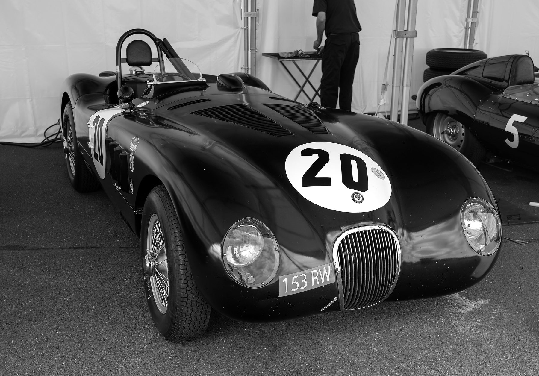 A dark sports-racing car numbered 20, registration 153 RW, stands in a paddock tent; behind it a man, his head out of the frame, works at a folding table, and a second sports car is parked at the right.