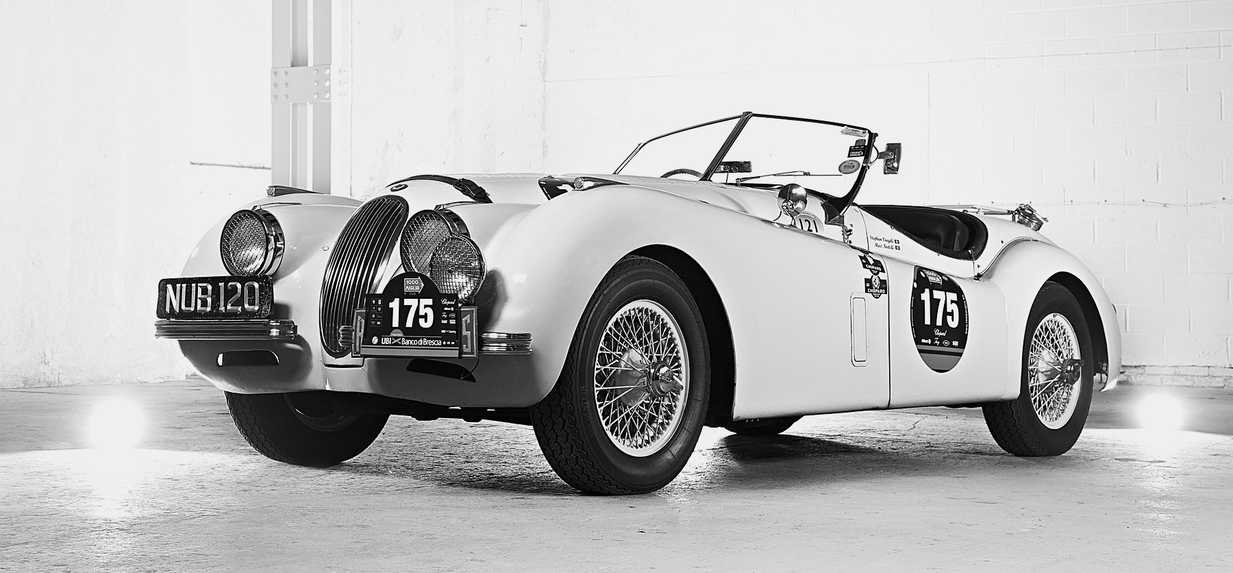 A pale open two-seater with wire wheels, spot lamps and rally plates numbered 175 stands in a whitewashed studio, lamps glowing at floor level.