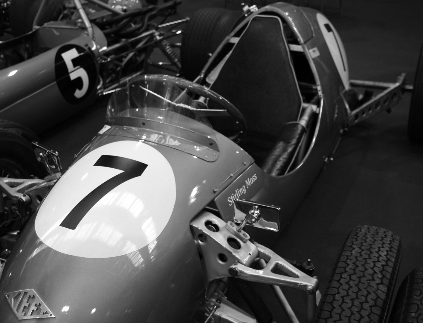 A low open single-seater with a large 7 in a white disc on its nose, a name in script beside the cockpit and a diamond badge, among other racing cars at a show.