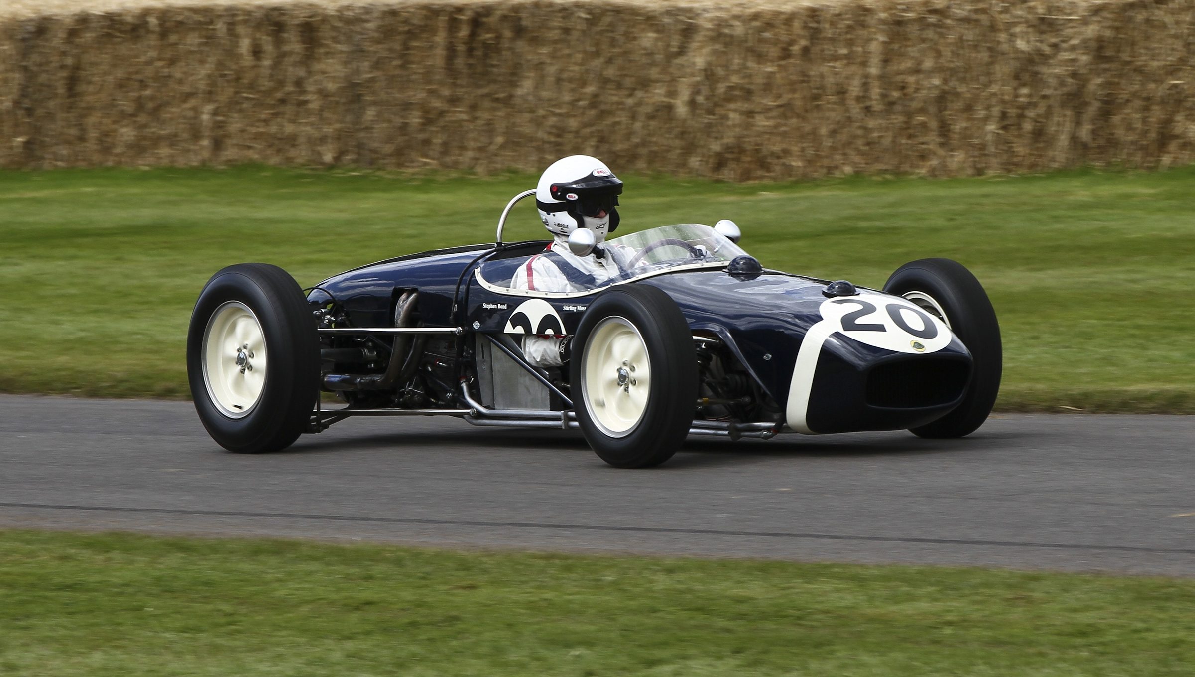 A small dark-blue rear-engined racing car with a white band, numbered 20, runs at speed between grass verges and straw bales; its driver wears a white helmet.