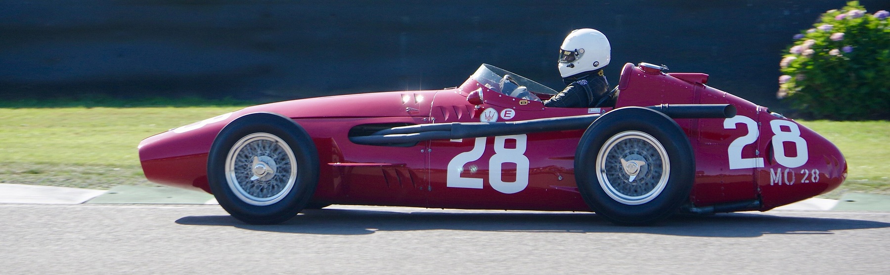 A red front-engined racing car numbered 28 on its nose and flank goes by at speed in front of a dark wall, its driver in a white helmet; flowering shrubs grow at the right.