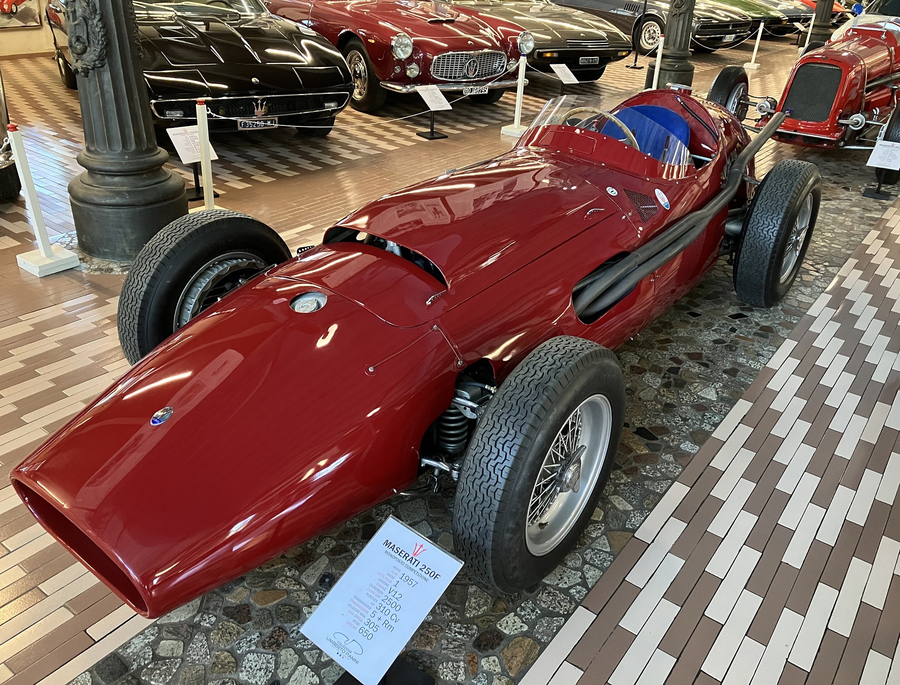 A long red front-engined racing car with wire wheels and exhaust pipes along its flank stands on a patterned floor, a placard before its nose, road cars behind it.