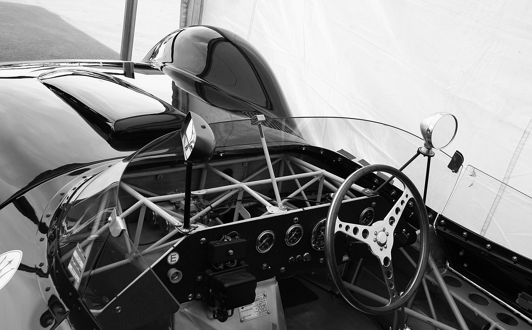 Looking down into the cockpit of an open sports-racing car: a dark-rimmed steering wheel with drilled spokes, a dashboard of round dials, and behind it a lattice of thin pale tubes forming the frame, under a curved windscreen.