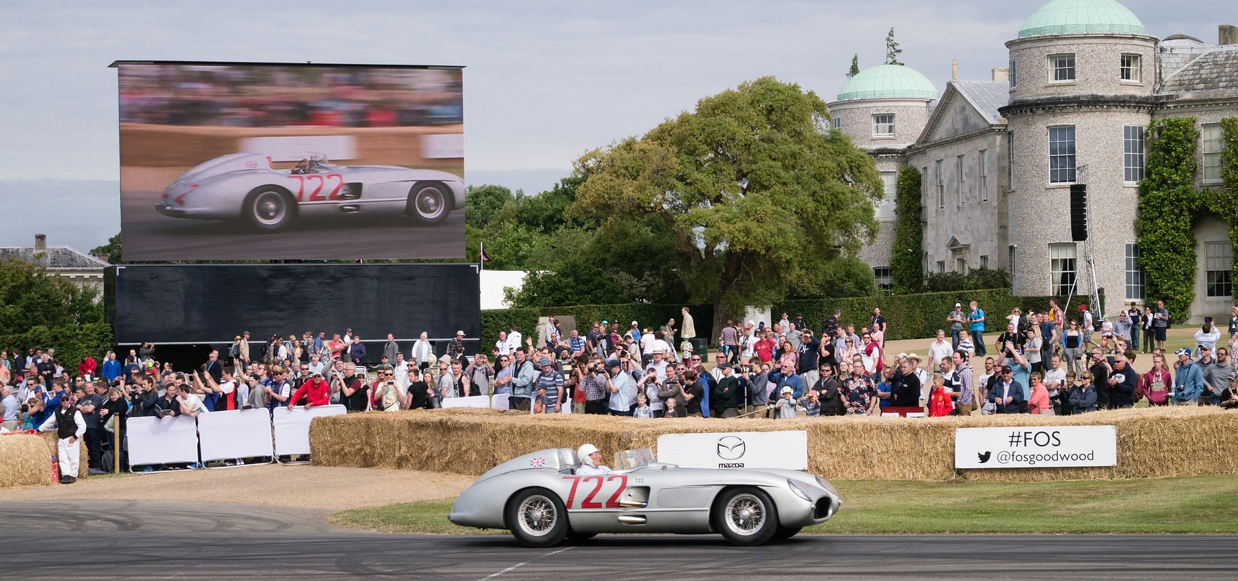 A low silver open sports-racing car numbered 722 in red runs past straw bales and a large crowd in front of a country house; a big screen above the crowd shows a silver car numbered 722.