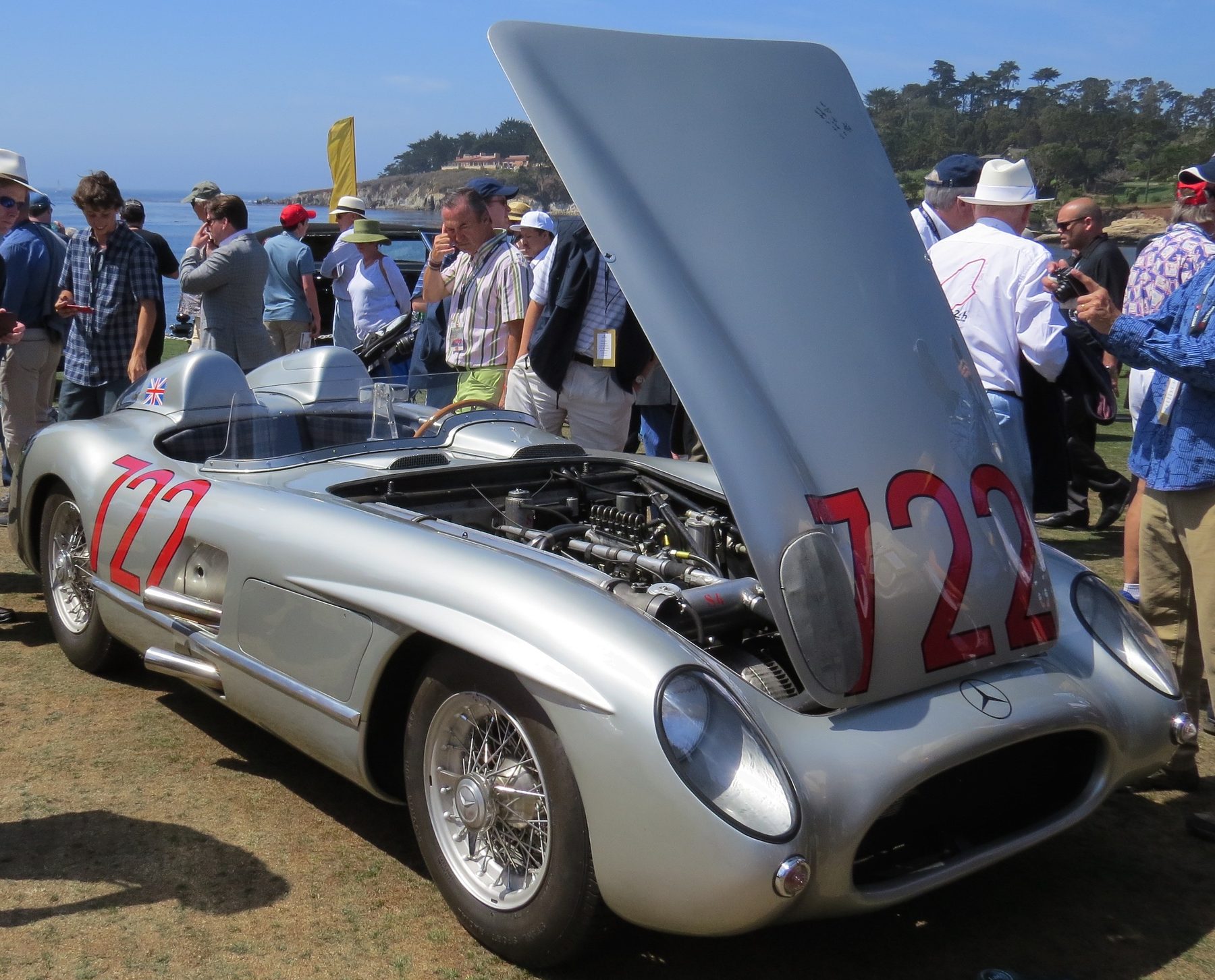 A long silver open sports-racing car with 722 painted in red on its flanks and bonnet stands on a lawn by the sea, the bonnet raised over its engine; visitors in sun hats crowd round it.