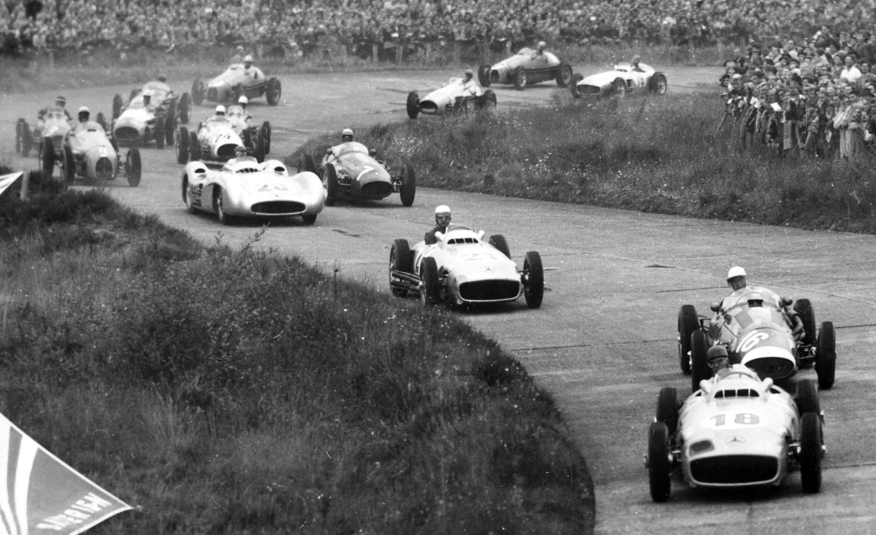 Racing cars stream round a curving bend between a grass bank and a hillside packed with spectators; an open-wheeled car numbered 18 leads another numbered 16, whose driver wears a white helmet, and a streamlined car and a long line of others follow; a striped pennant hangs into the lower left corner.
