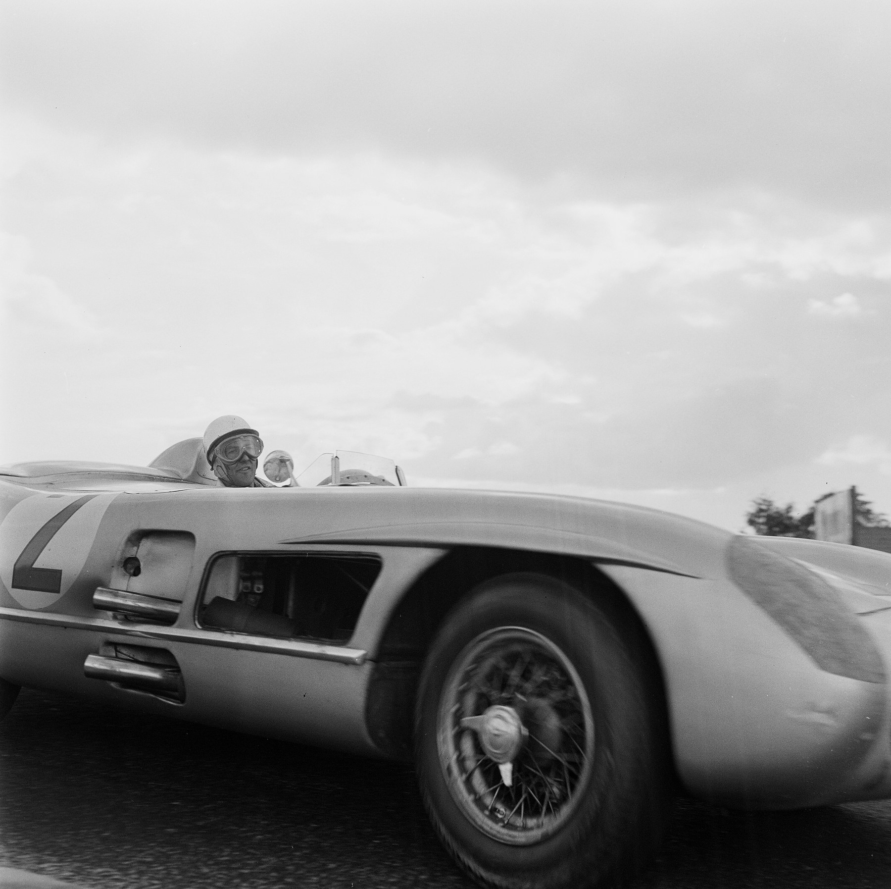 A low view of a streamlined open sports-racing car at speed, its front wheel blurred, a driver in a white helmet and goggles in the cockpit, a wide clouded sky above.