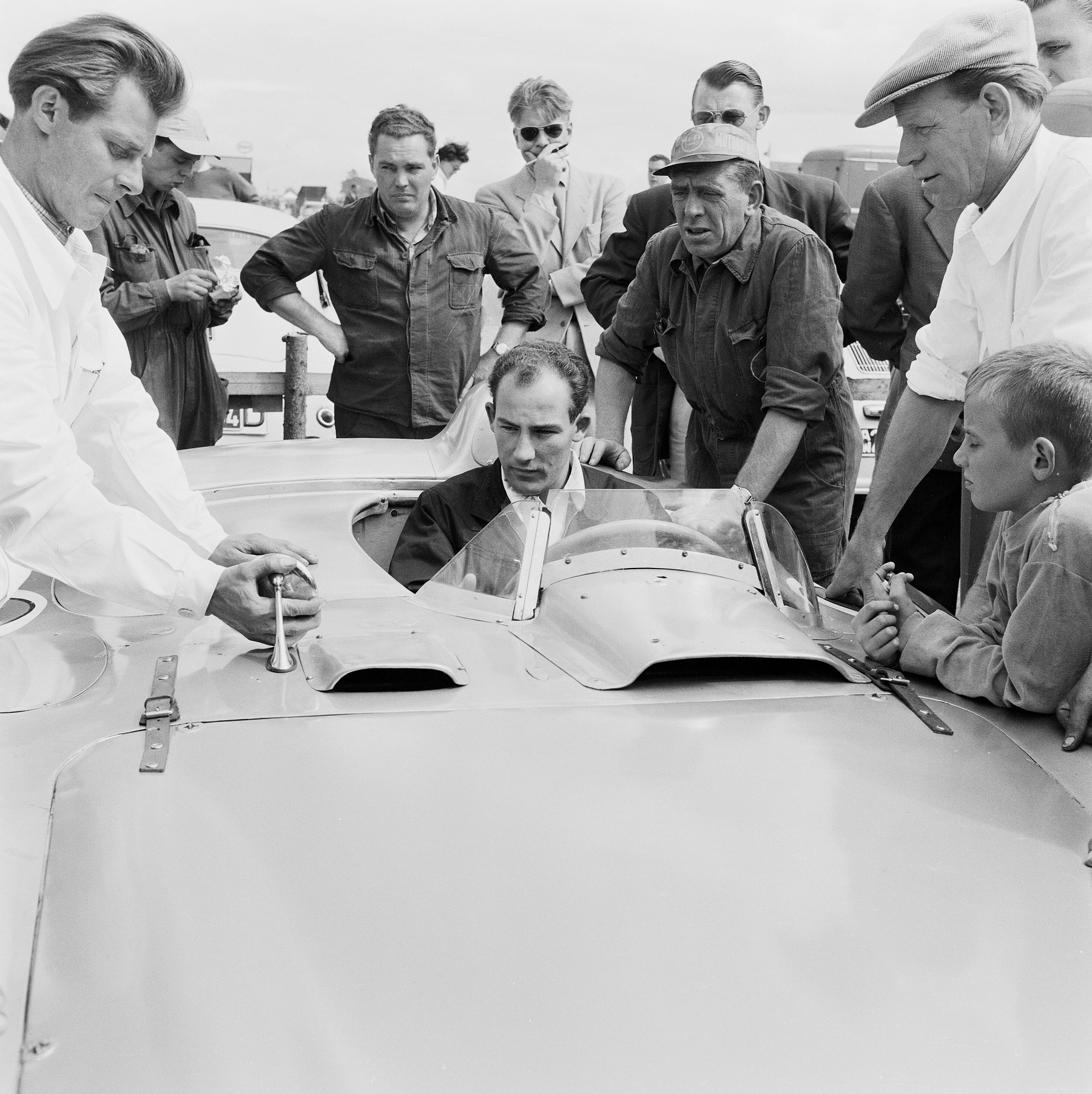 A young man in a dark jacket and an open-necked shirt sits in the cockpit of a long, low sports-racing car, looking ahead; eight men stand round it, one in a white coat with his hand on a mirror on the bodywork, and a boy rests his arms on the car.