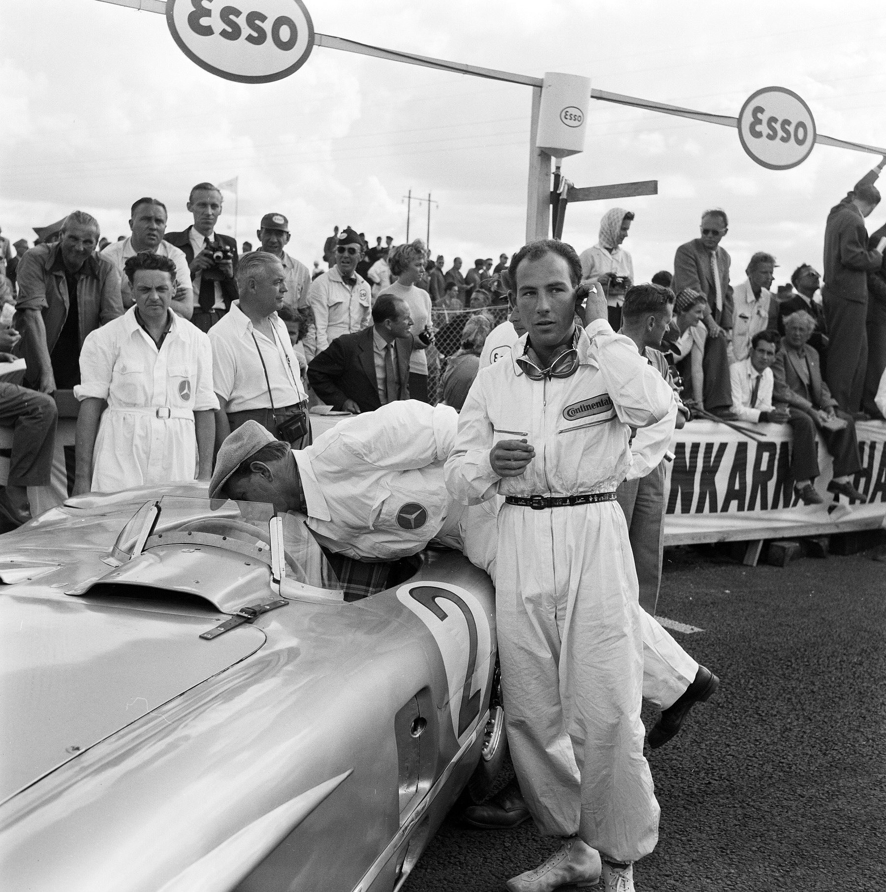 A young man in white overalls, goggles round his neck and one hand at his ear, stands beside a sports-racing car numbered 2; a mechanic leans into its cockpit, and spectators and photographers crowd a barrier beneath round petrol-company signs.