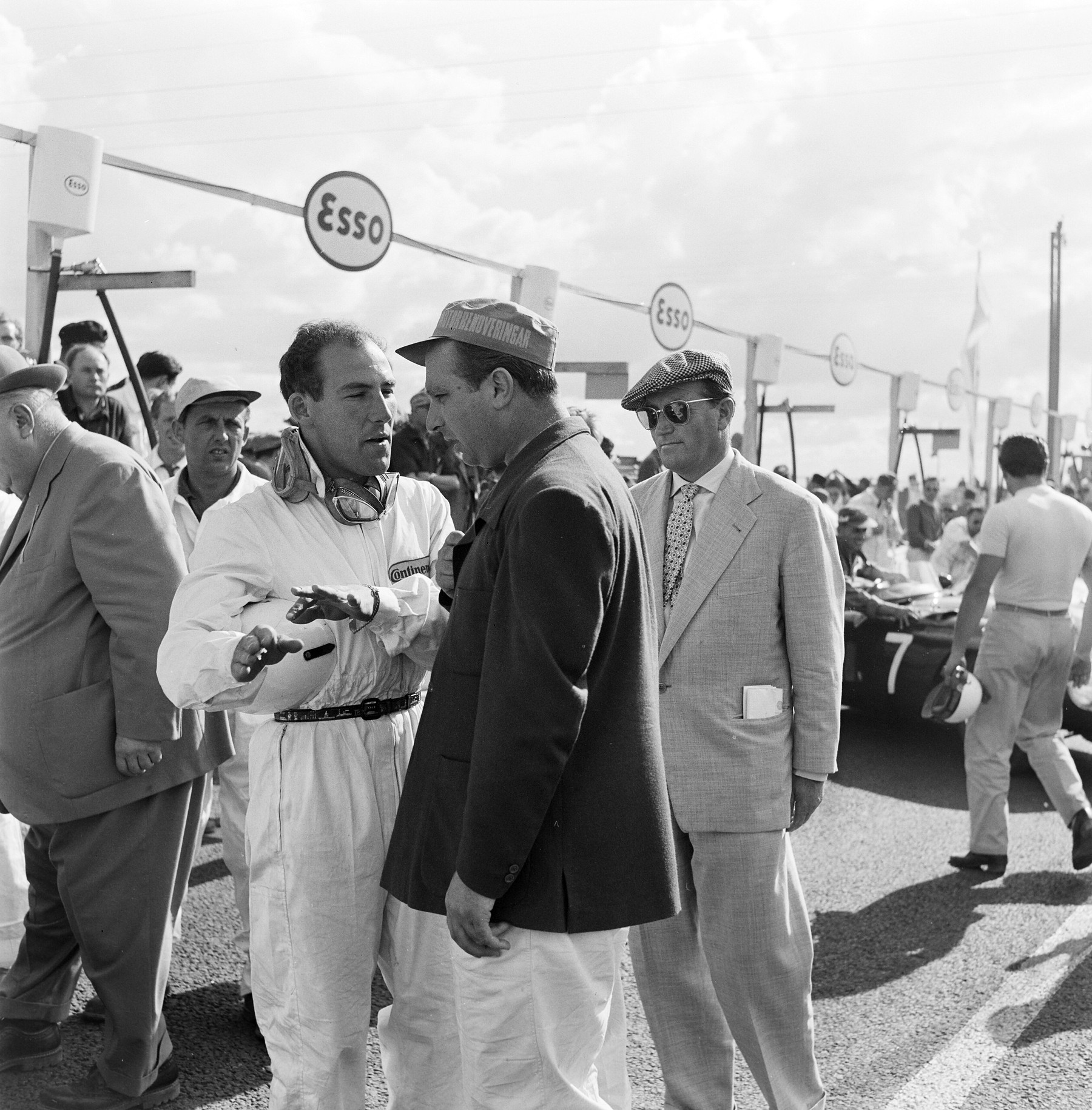 A young man in white overalls, goggles round his neck and a helmet in his hand, gestures as he talks to a man in a dark jacket and peaked cap; a man in a pale suit and sunglasses looks on, and others stand in the pit road behind.