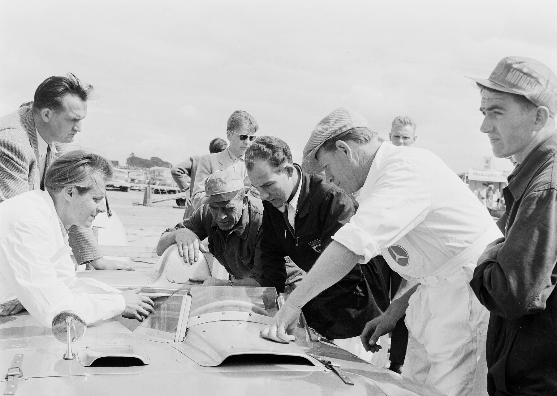 A young man in a dark zipped jacket, a shield-shaped badge with a Union Jack on its breast, leans in over the cockpit of a smooth open sports car with two men in white overalls, one wearing a cap; more men in caps and jackets crowd round, and parked cars stand in the distance.