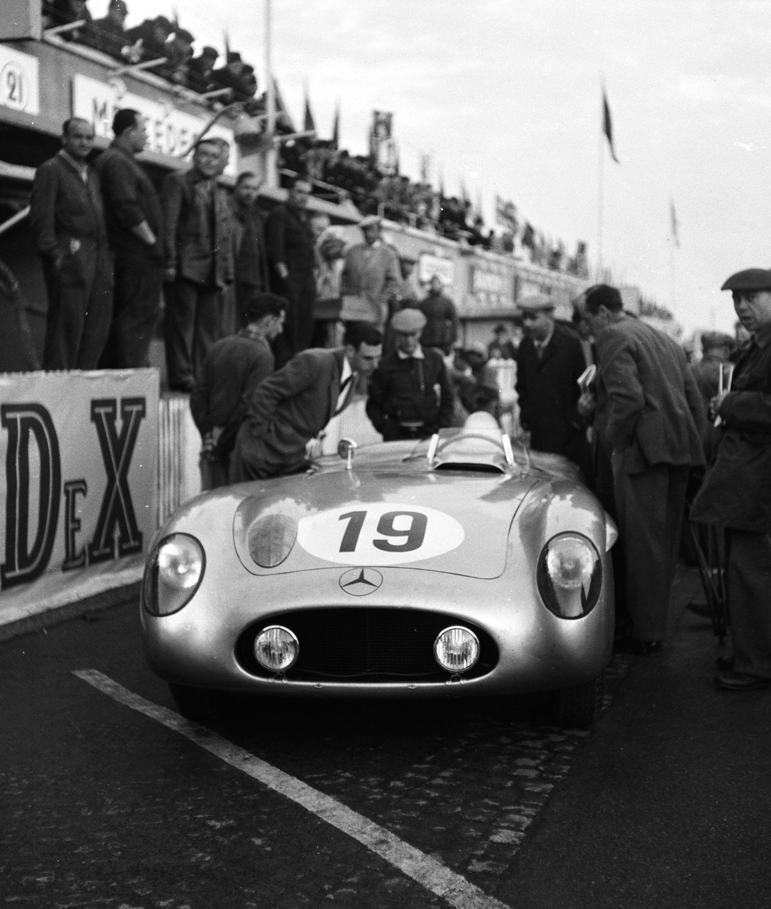 Nose-on at a pit, a low open sports-racing car numbered 19, a three-pointed star on its nose; men in caps and coats lean over it or stand round it, mechanics stand at the pit counter above, and people crowd a balcony behind.