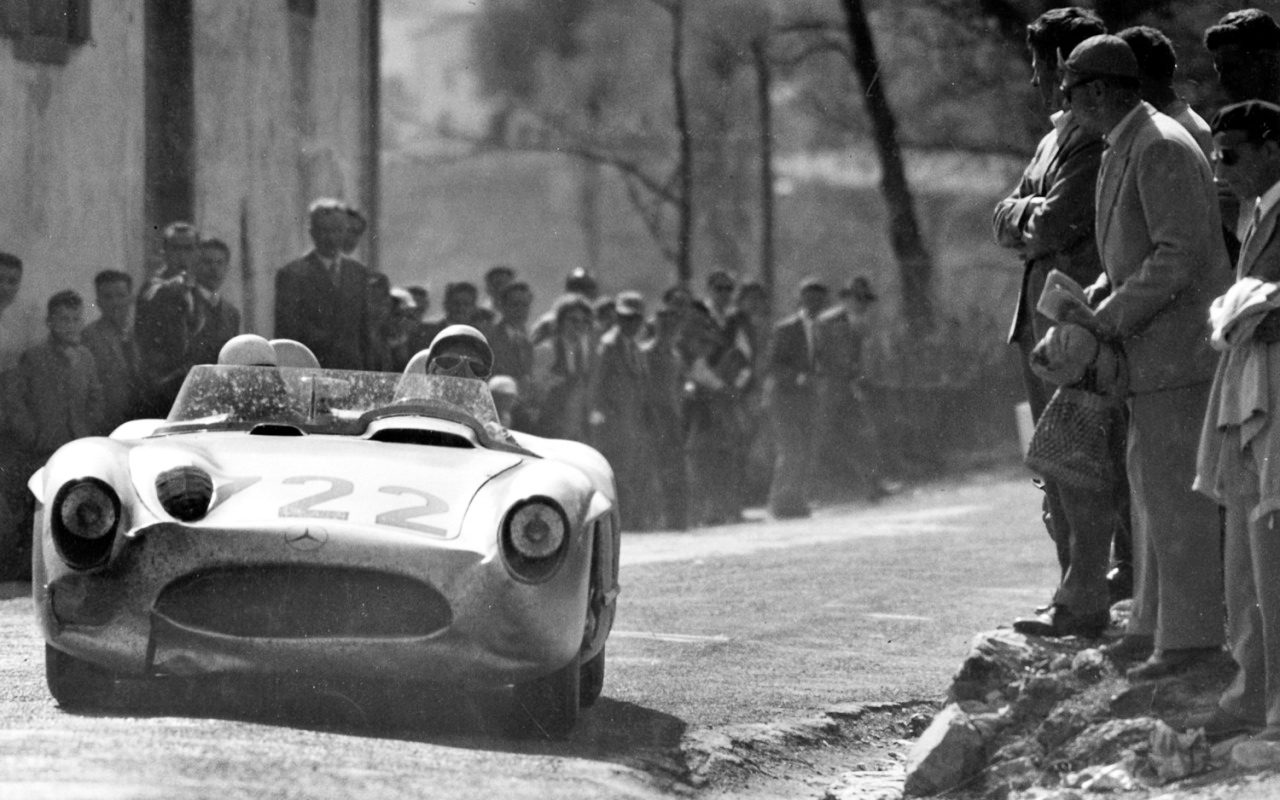 A dust-streaked open sports-racing car numbered 722 comes towards the camera on a village road, a helmeted driver in goggles behind its screen; men in coats and caps line the road, some standing on rocks at the right.