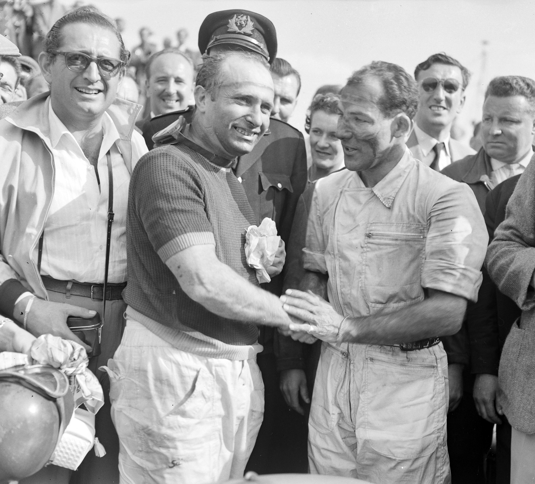 Two drivers shake hands in a smiling crowd: an older man in a short-sleeved knitted shirt and pale trousers, and a younger man in overalls, his face streaked with dirt; a man in a peaked cap stands behind them, and a man in glasses at the left holds a camera.
