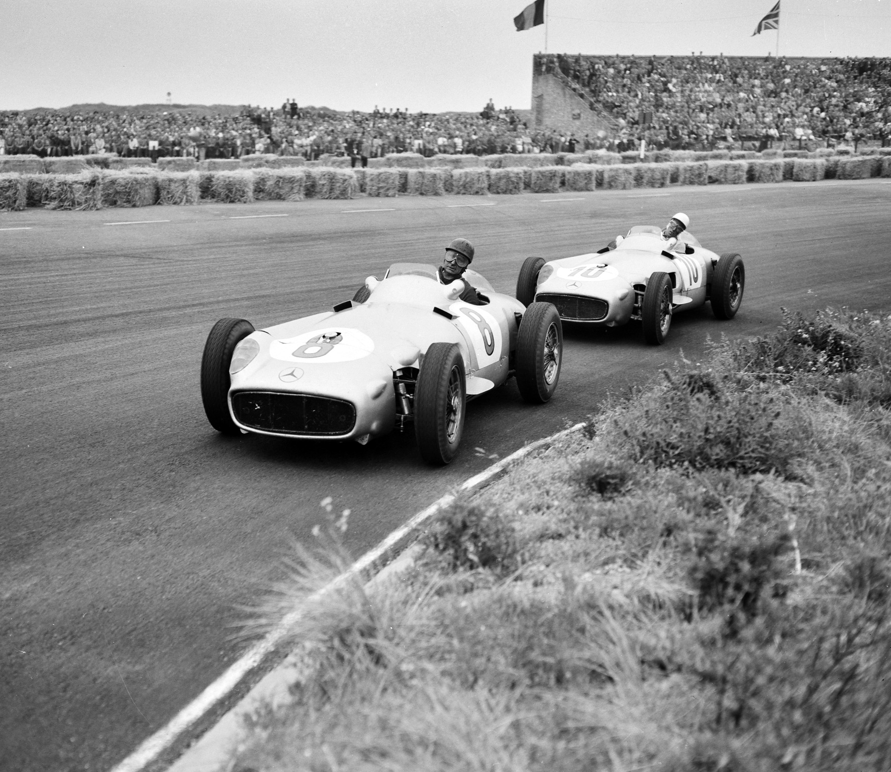 Two pale, smooth-bodied open-wheeled racing cars, numbered 8 and 10, run nose to tail along a track lined with straw bales below a packed grandstand flying flags; the first driver wears a dark helmet and goggles, the second a white helmet.