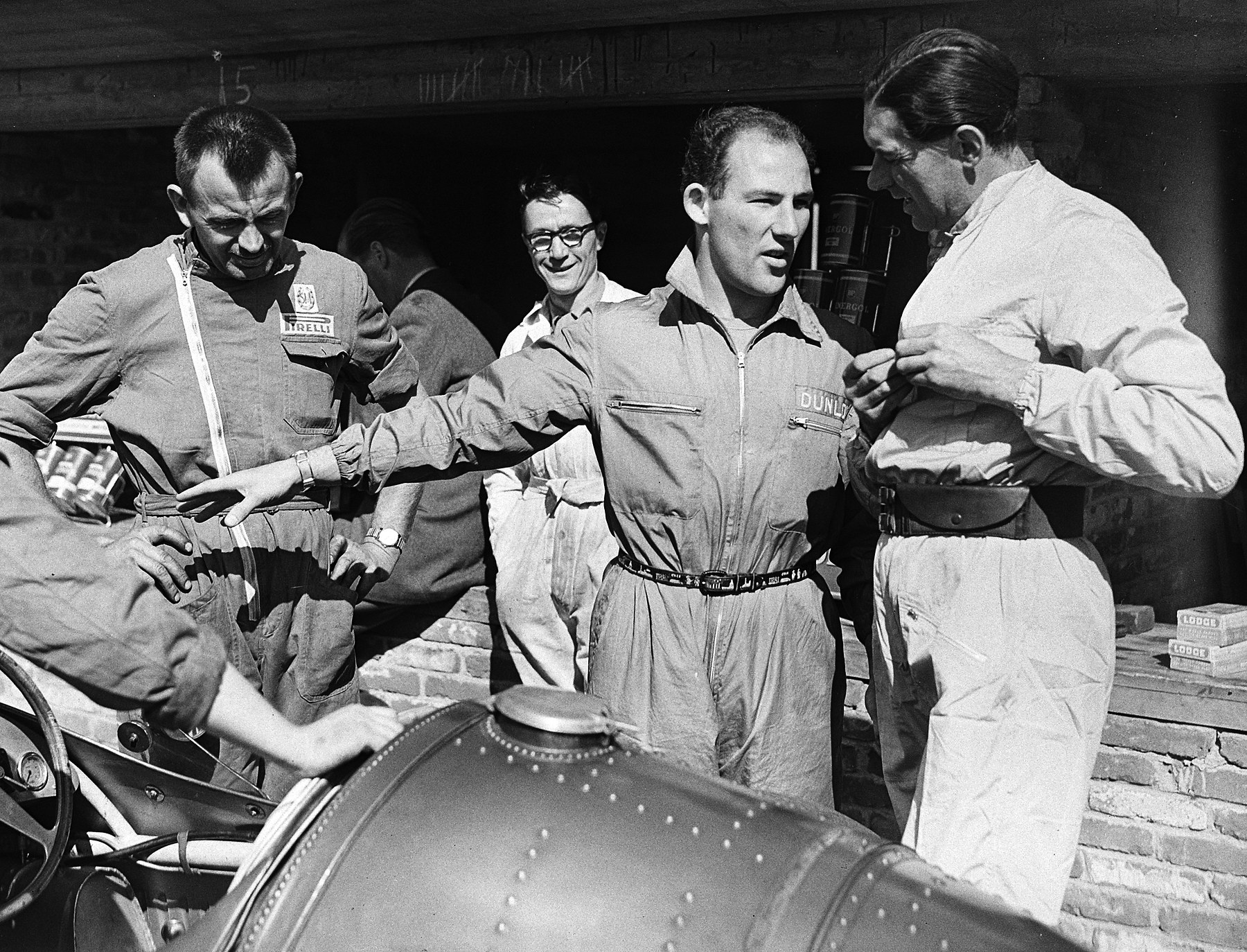 In front of a brick garage a young man in overalls stretches out one arm as he talks; a taller man in pale overalls stands at the right, two men in work clothes and a smiling man in glasses stand at the left and behind, and the riveted tail of a racing car fills the foreground.