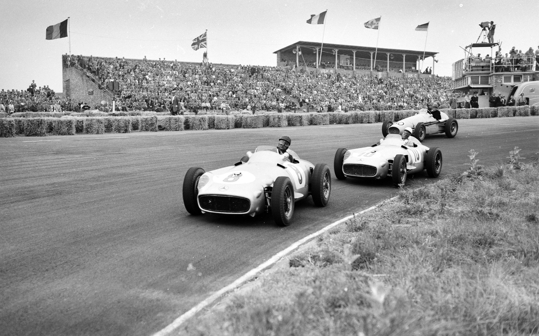 Two open-wheeled racing cars, numbered 8 and 10, sweep through a bend close together in front of a packed grandstand under flags; a third car follows on the outside, and a cameraman stands on a tower at the right.