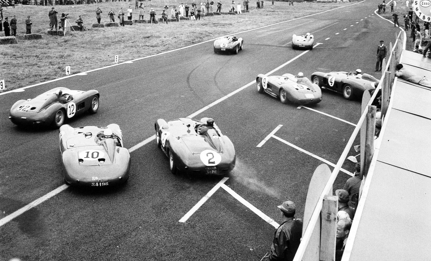 Seen from above, sports-racing cars pull away from the start beside a pit wall on a wide track, the nearest numbered 10, 2 and 12, with more ahead; photographers and marshals stand in the field beyond.