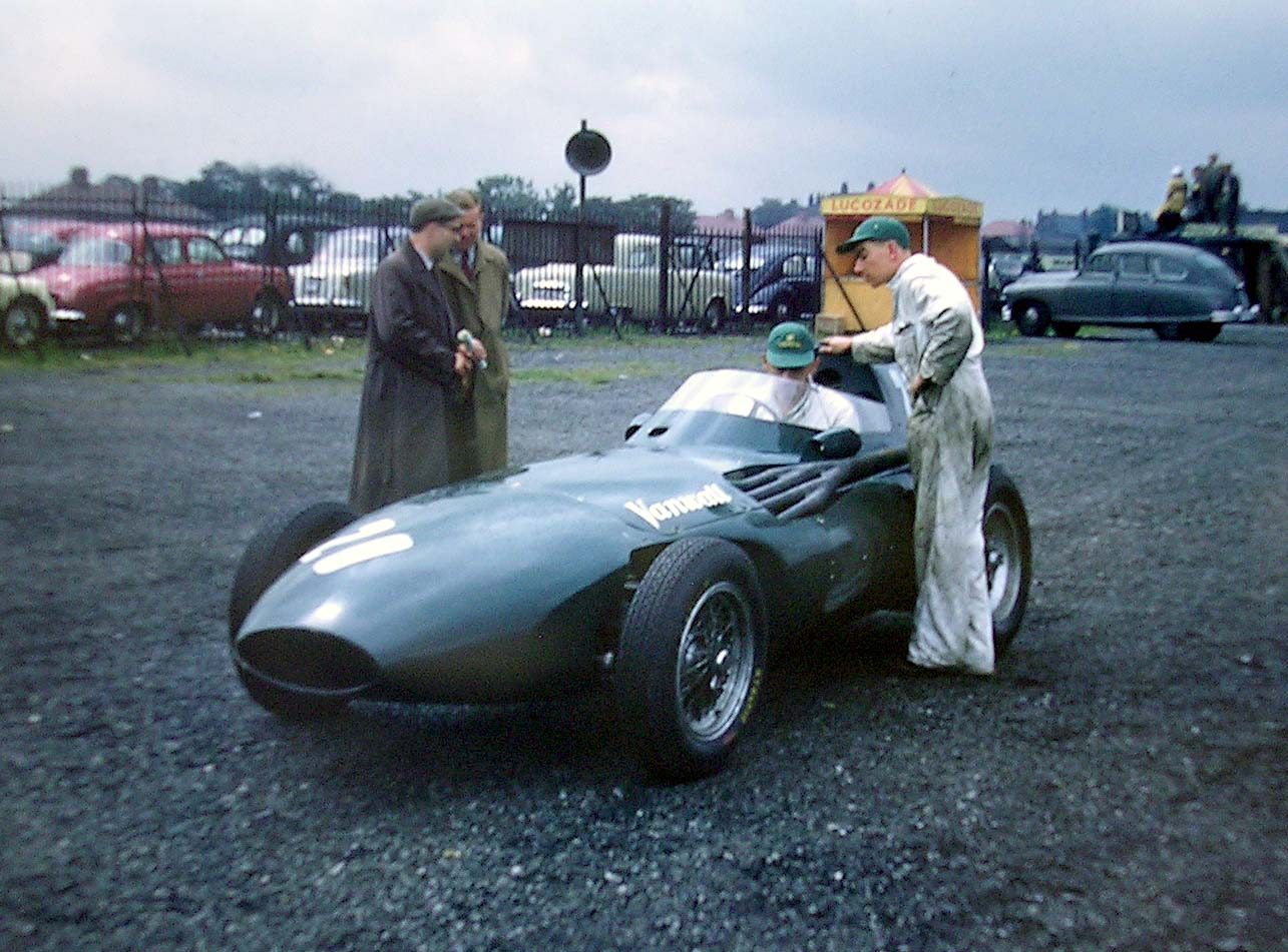 A low, streamlined front-engined racing car stands on cinders by a car park. A man in a cap sits in its cockpit, a mechanic in overalls stands at its side, and two men in raincoats stand by its nose.