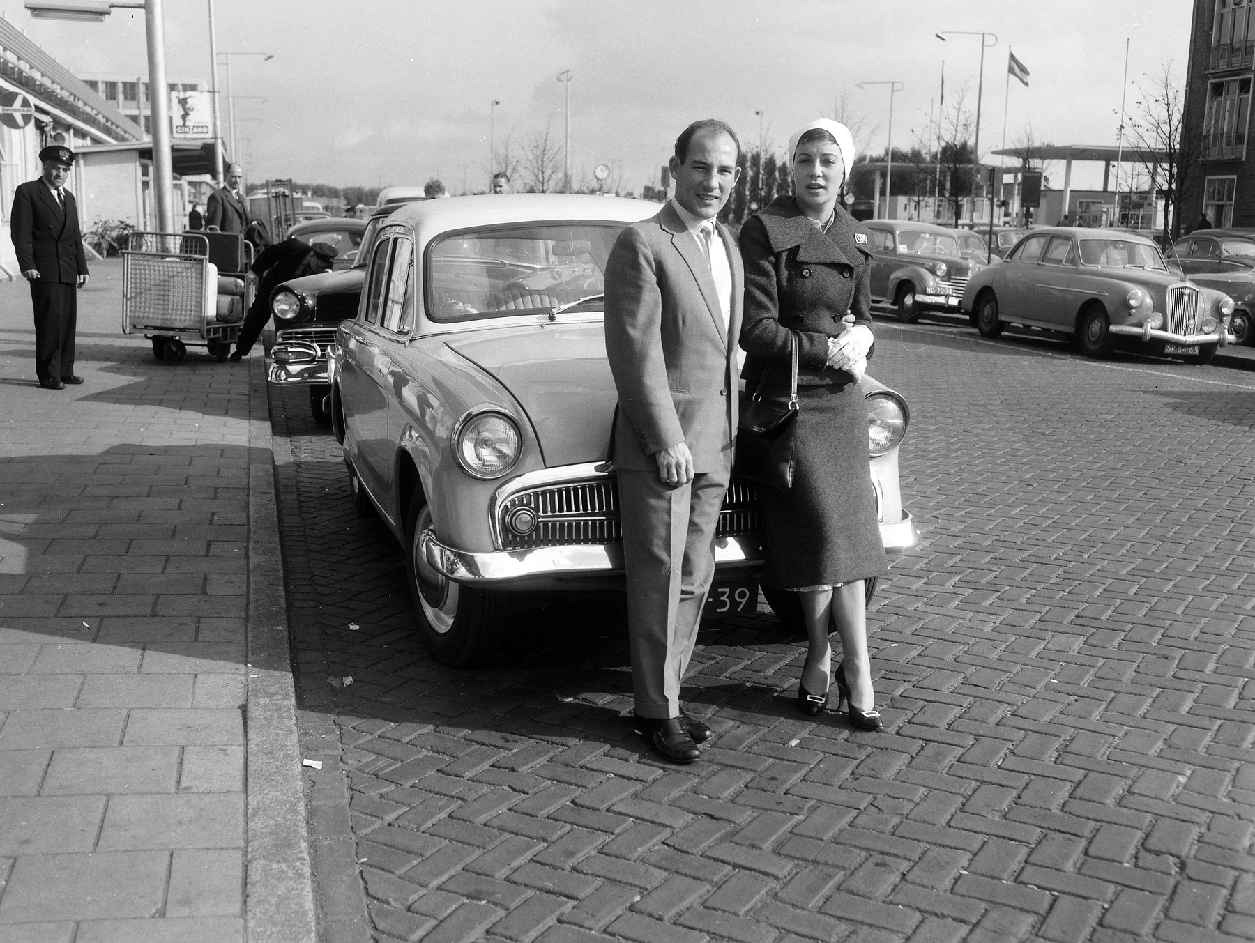 A man in a light suit and tie and a woman in a dark coat, a white headscarf and gloves stand side by side against the front of a parked saloon car on a cobbled forecourt outside an airport building; a uniformed porter stands on the pavement behind.
