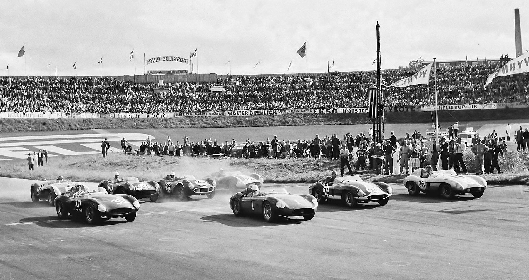 Open sports racing cars pull away from a start in a haze of dust, watched by people on a bank and a packed grandstand; the driver of the car in the middle, numbered 1, wears a white helmet.