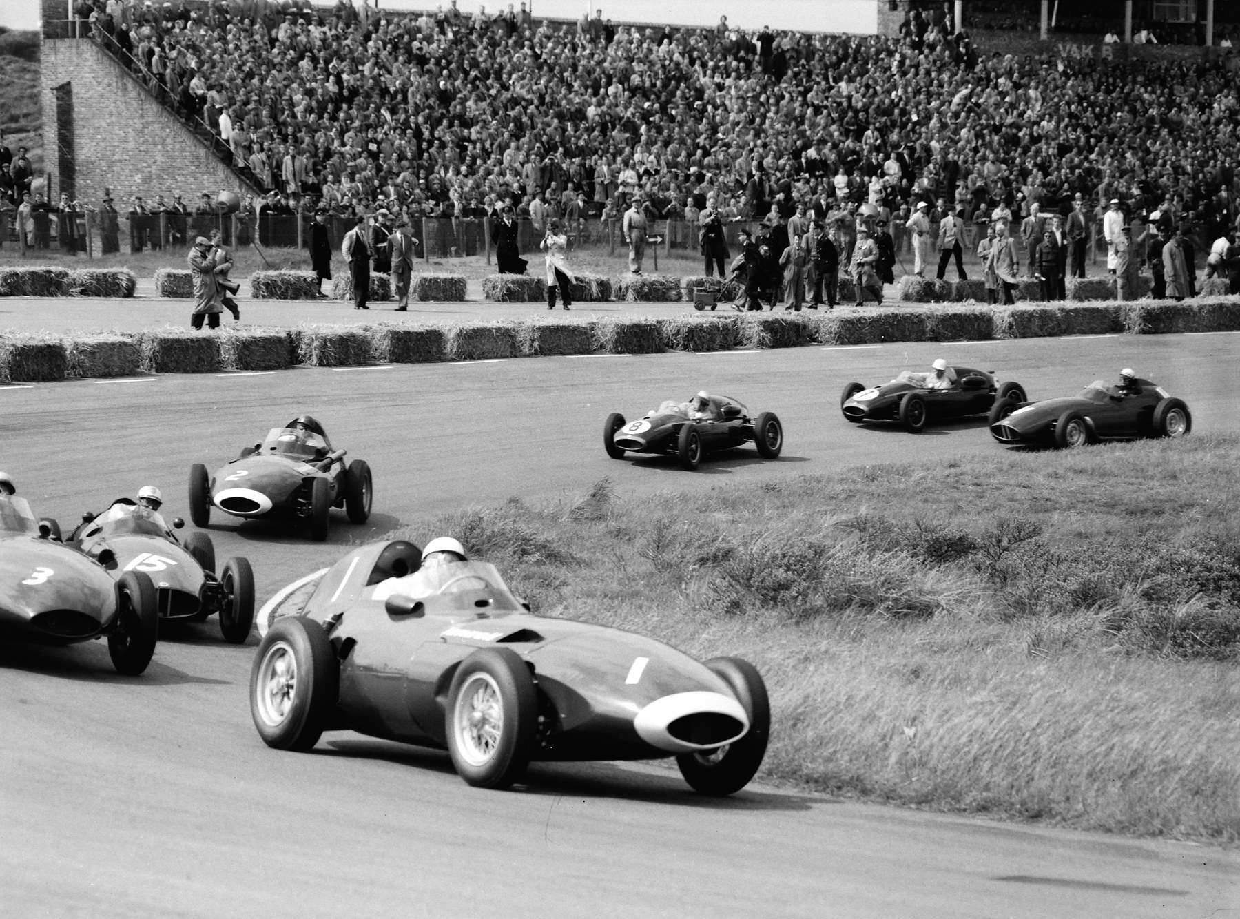 A streamlined racing car numbered 1, its driver in a white helmet, leads a line of racing cars through a bend between a grass bank and a crowded stand.