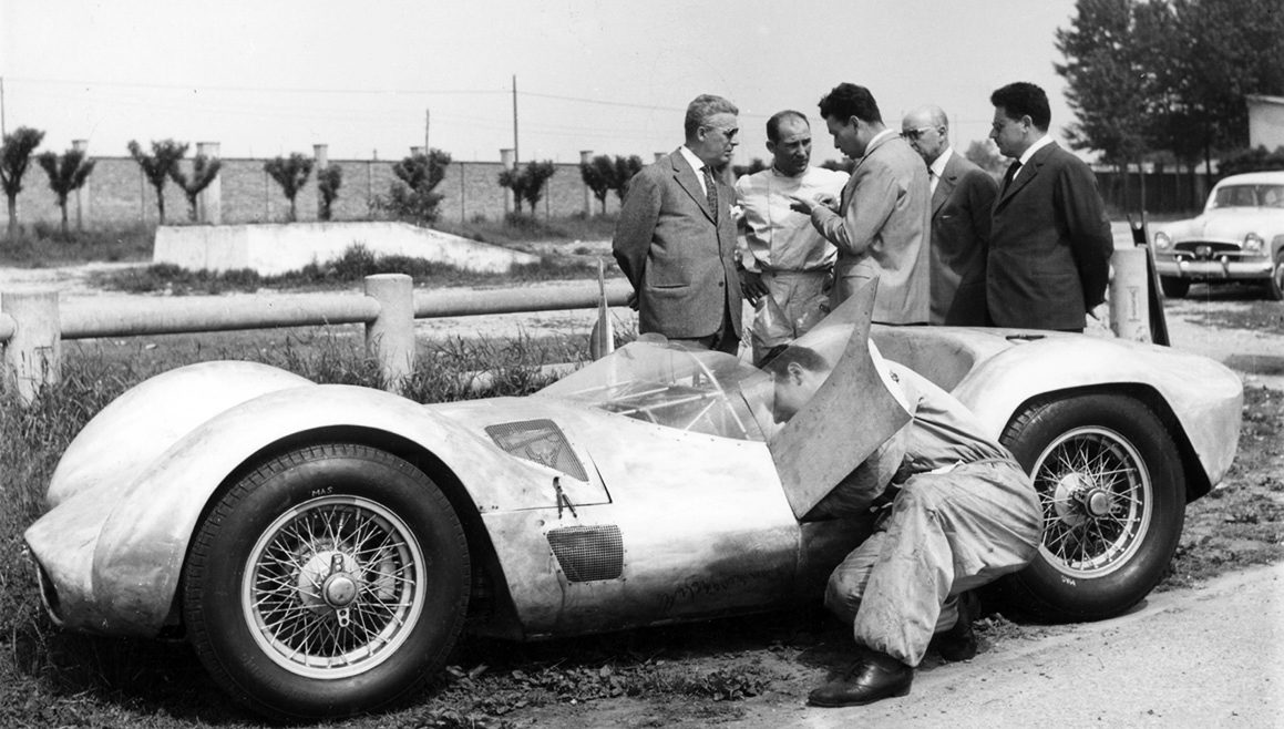 A man in pale overalls stands among four men in suits beside an unpainted open sports-racing car while a mechanic in overalls leans into its cockpit; trees and a wall behind.