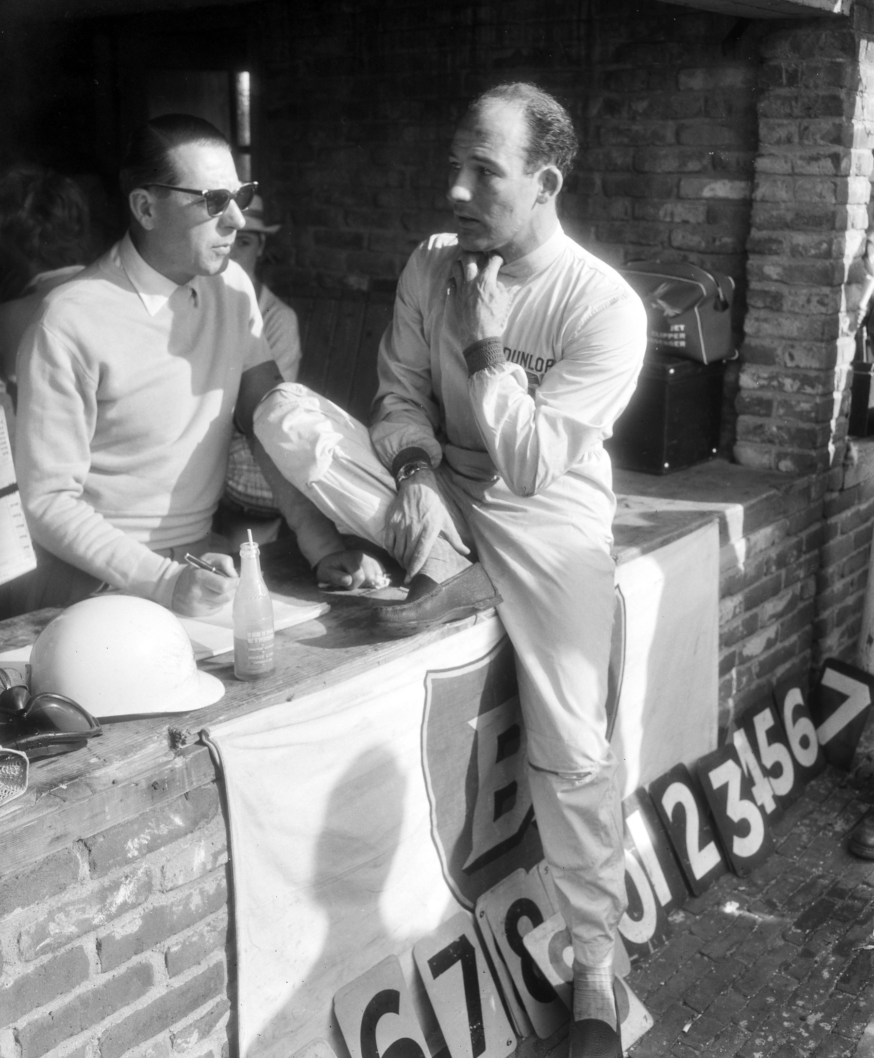 A man in pale overalls sits on a brick pit counter, one hand at his chin, talking to a man in sunglasses who writes on a pad; a helmet lies on the counter and pit-board numerals lean against the wall below.