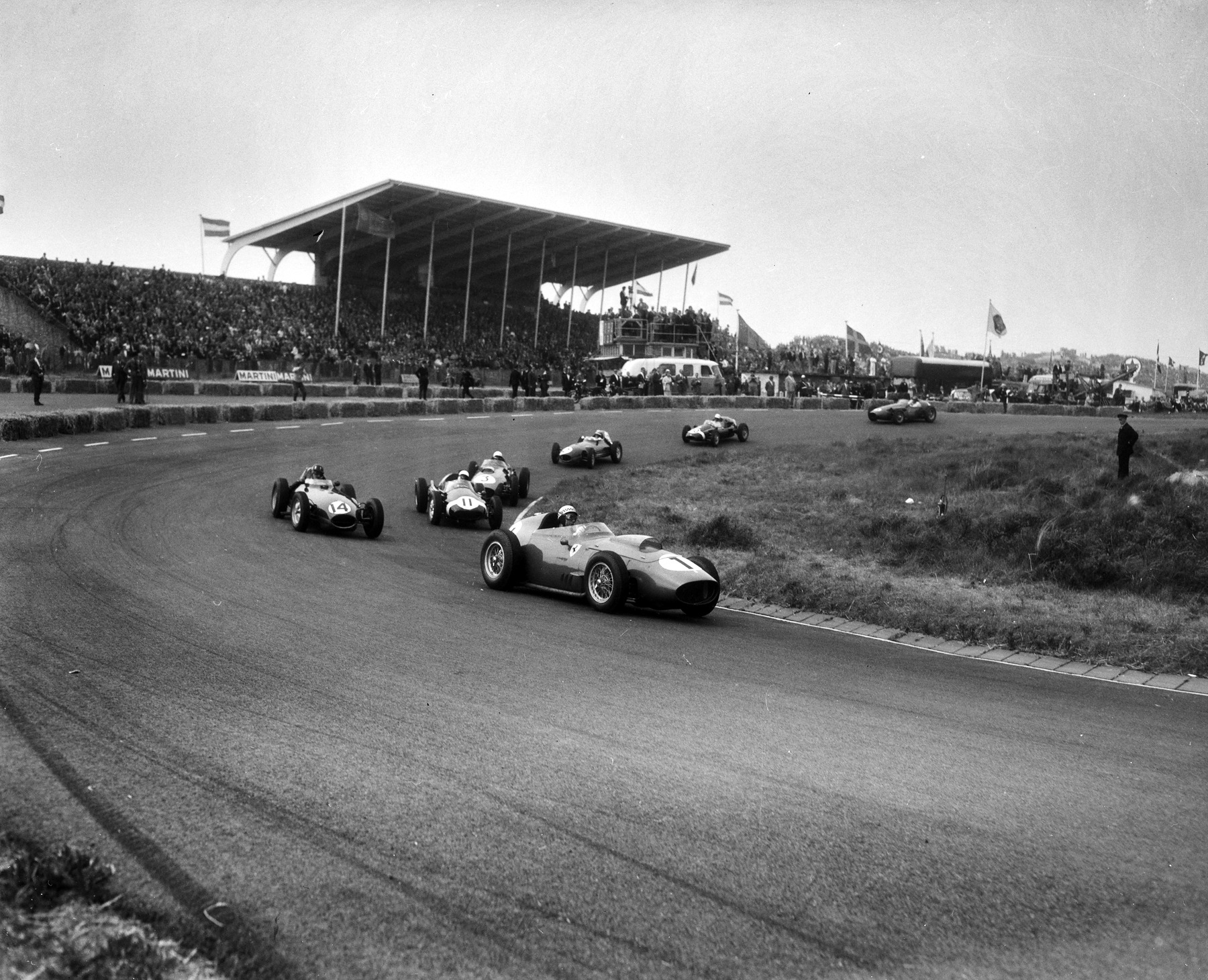 Racing cars sweep into a bend below a large covered grandstand flying flags: a front-engined car numbered 1 leads; cars numbered 3, 11 and 14 follow, and the rest of the field strings out behind.