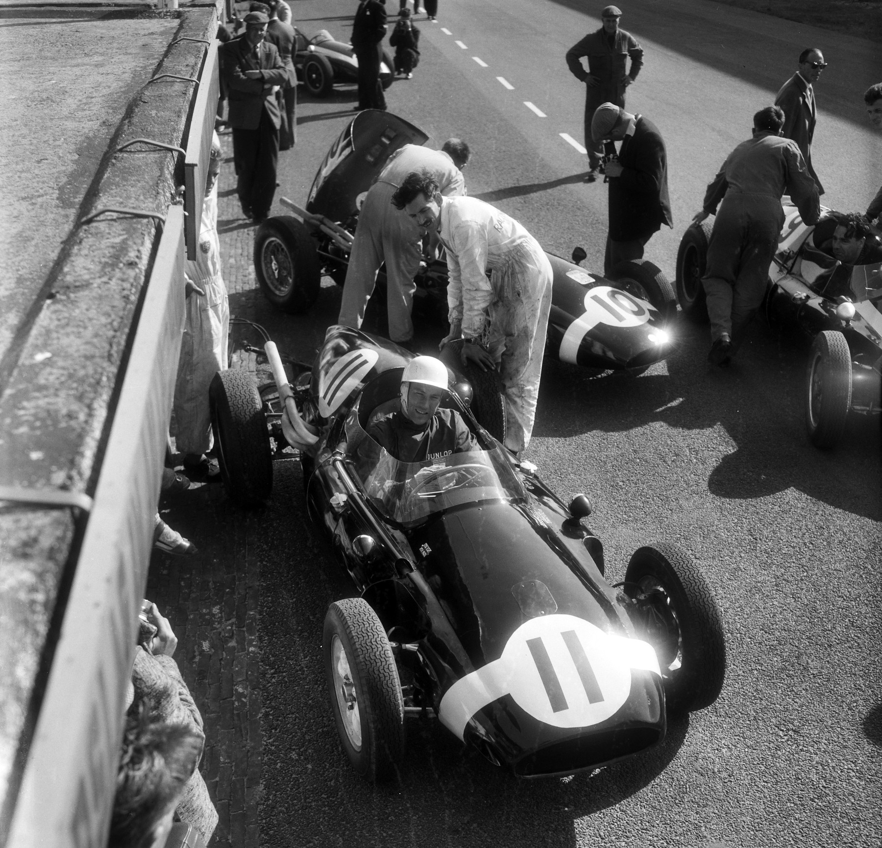 Seen from above, a smiling driver in a white helmet sits in a small rear-engined racing car numbered 11 beside a pit wall; men in overalls lean over another car, numbered 10, alongside, and others stand in the pit lane beyond.