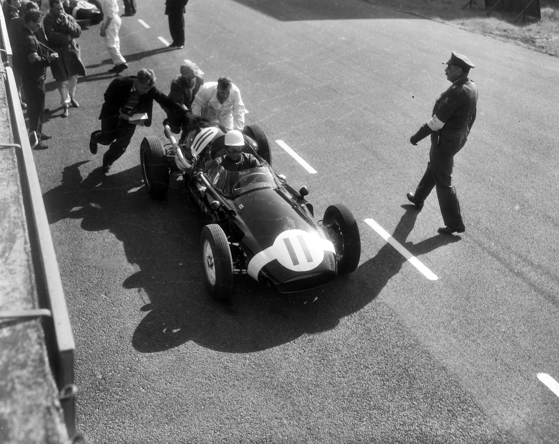 Seen from above, three men push a small rear-engined racing car numbered 11 away from a pit wall; its driver wears a white helmet, a uniformed official walks towards it, and onlookers in coats stand along the wall.
