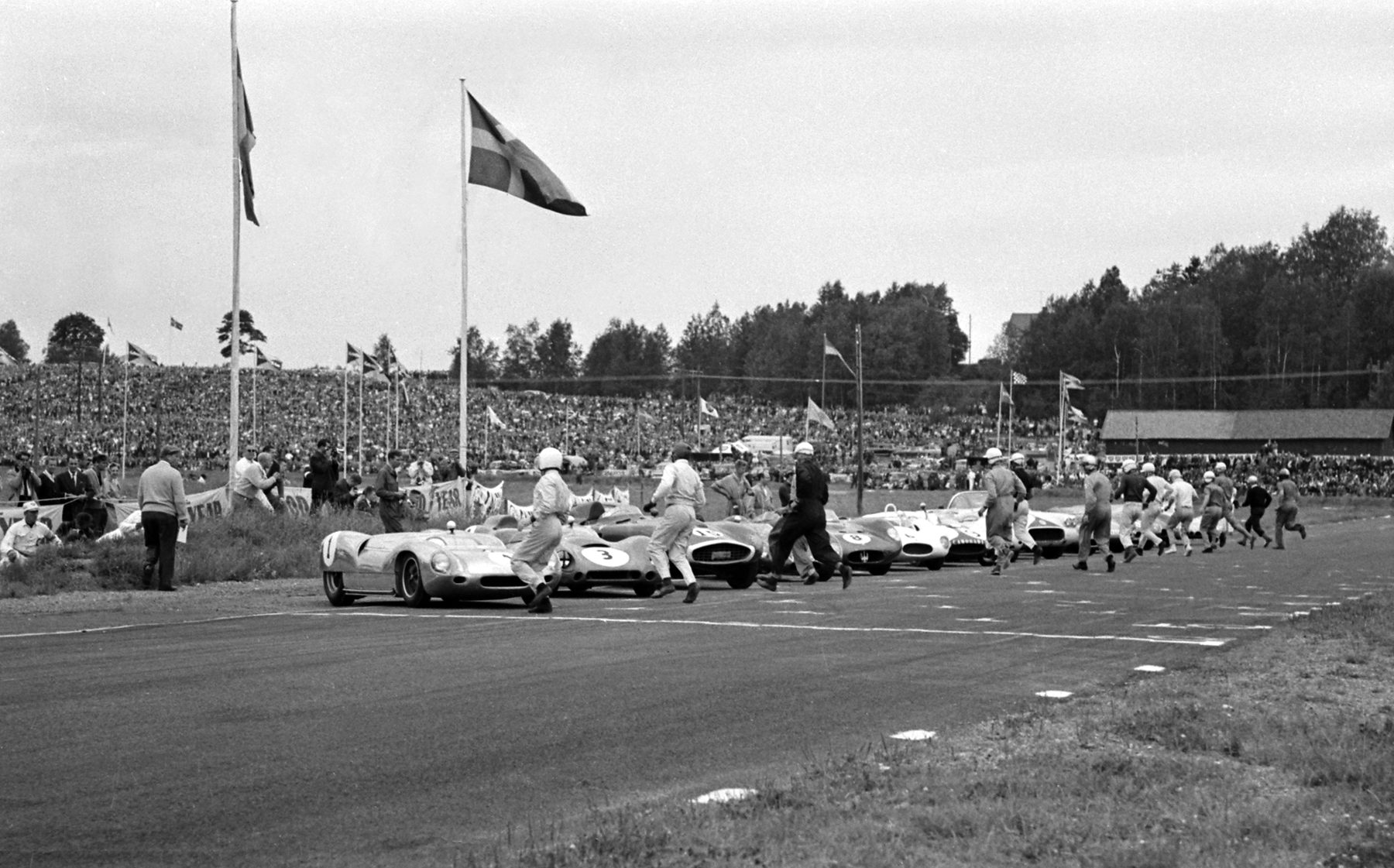 Drivers in white helmets and overalls run across a track to low sports racing cars lined up along the edge, flags on tall poles overhead and a huge crowd on the bank behind.