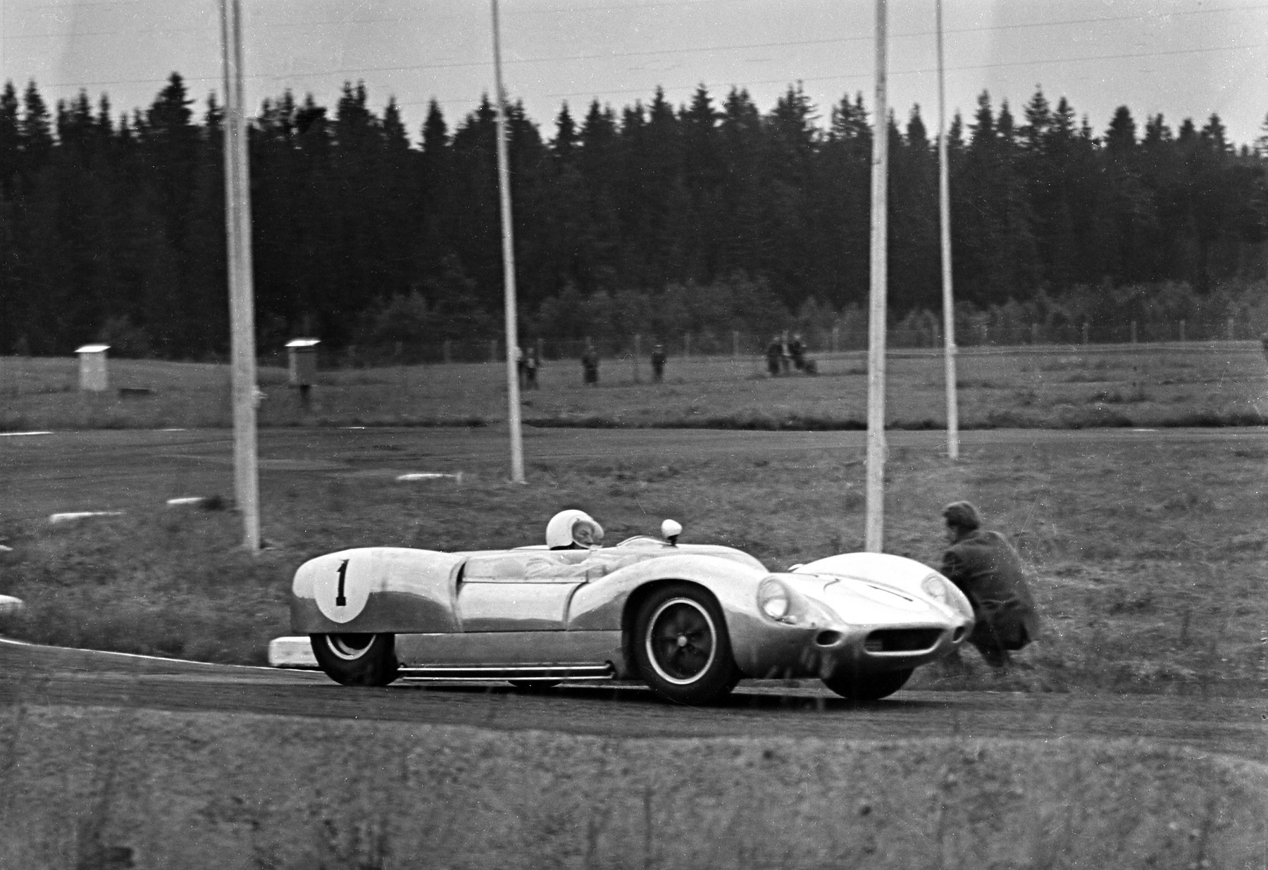 A low open sports racing car numbered 1, its driver in a white helmet, corners past a crouching man; telegraph poles stand along the track, a few spectators watch from the field and dark pine forest rises behind.