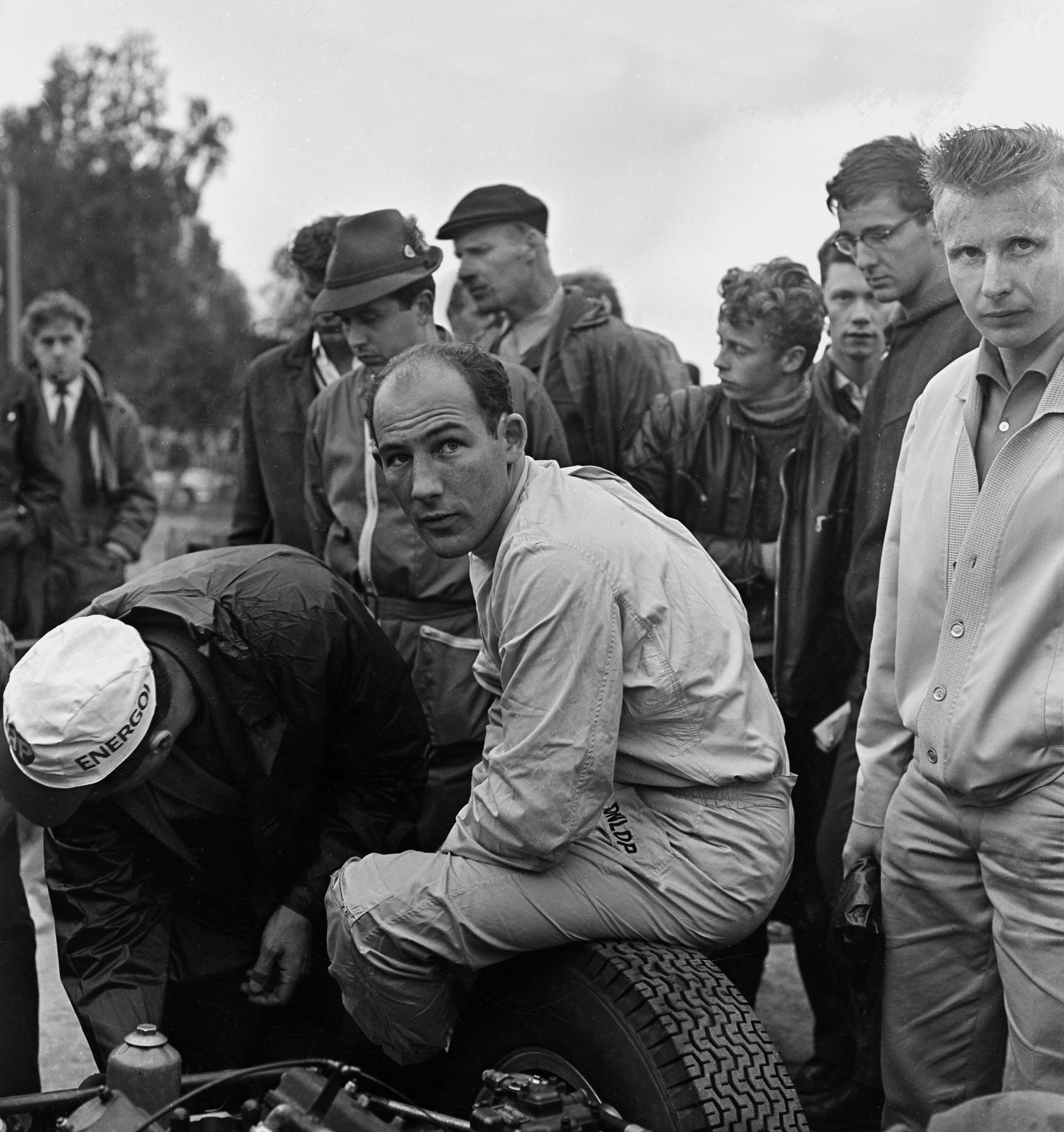 Moss, in pale racing overalls, sits on a rear tyre of a sports racing car and looks over his shoulder at the camera; a mechanic in a cap bends over the engine beside him, and a crowd of men and boys stands close behind.