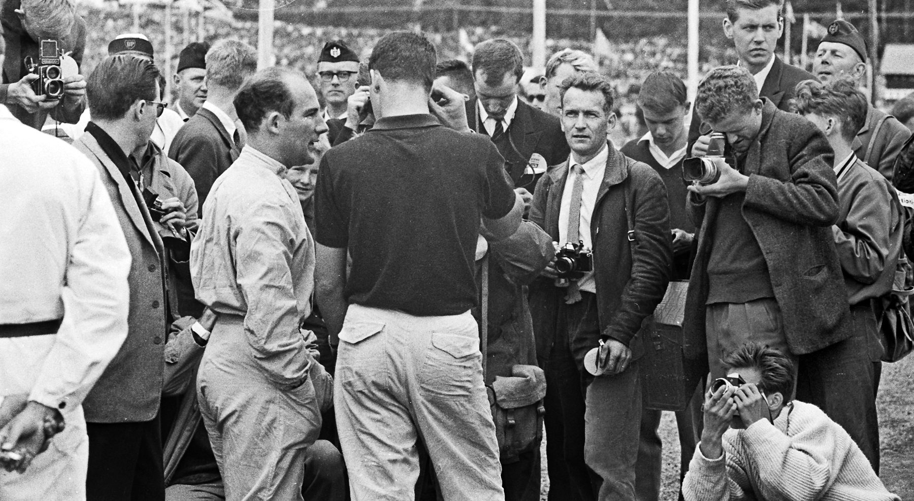 A press scrum: a man in pale overalls talks to a man seen from behind in a dark shirt, while photographers with cameras crowd round them, one kneeling in front.