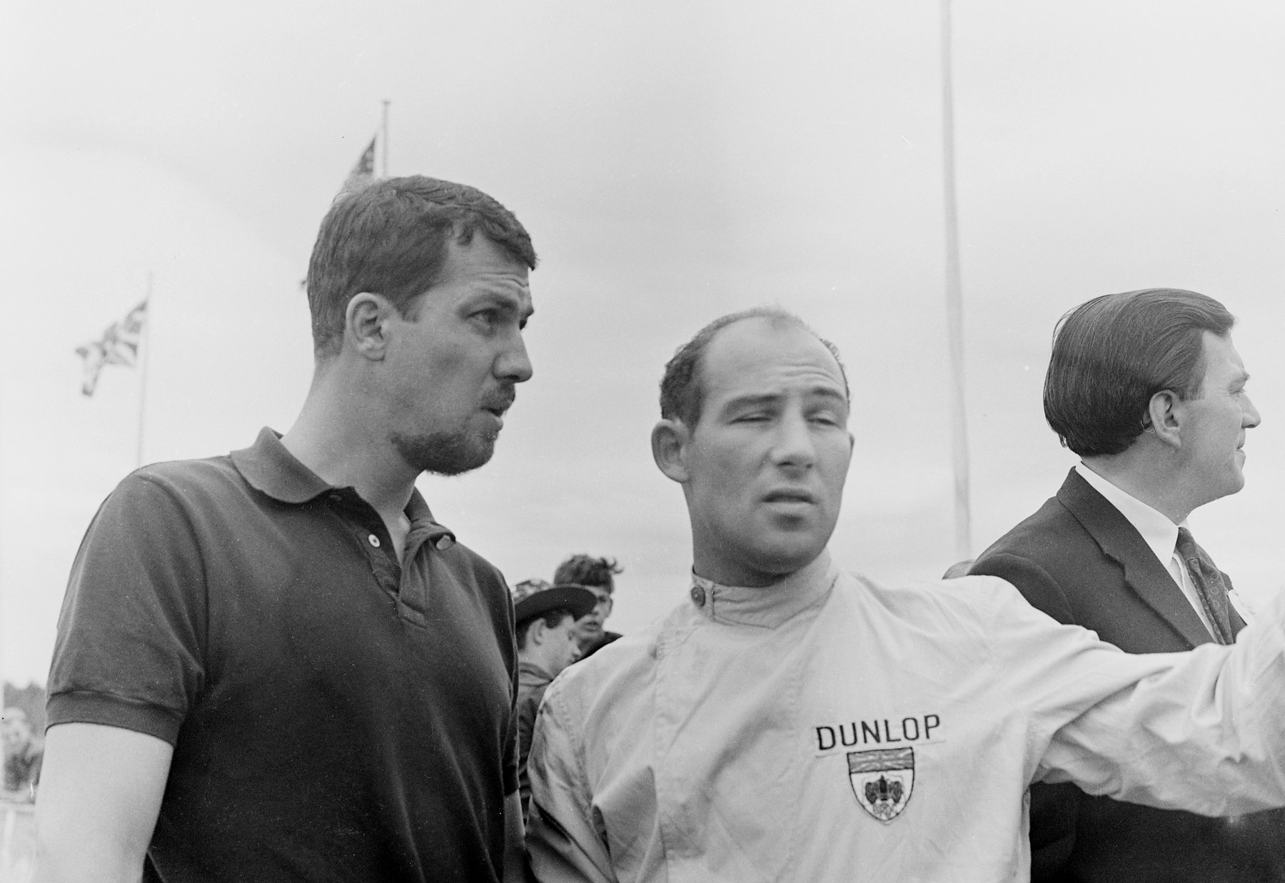 A bearded man in a dark polo shirt and a man in pale overalls with a badge on the chest stand side by side, looking off to one side, the second with an arm stretched out; a man in a suit stands at the right, and flags fly against a clouded sky.