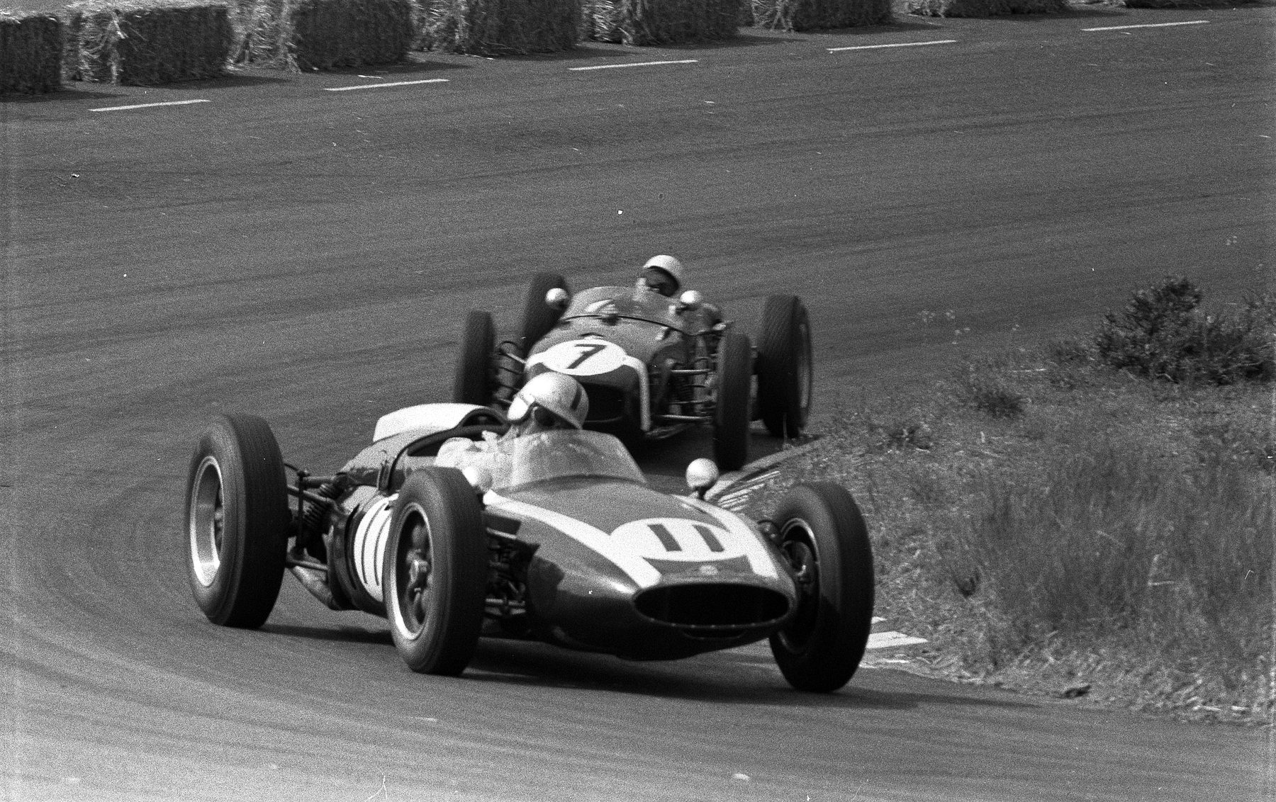 Two small rear-engined racing cars take a bend between straw bales and a grass verge, both drivers in white helmets; the nearer car is numbered 11, the one close behind it 7.