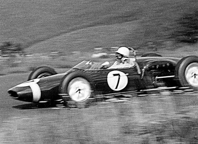 A panned photograph: a small open-wheeled racing car numbered 7, its driver in a white helmet, sharp against a blurred grass bank.