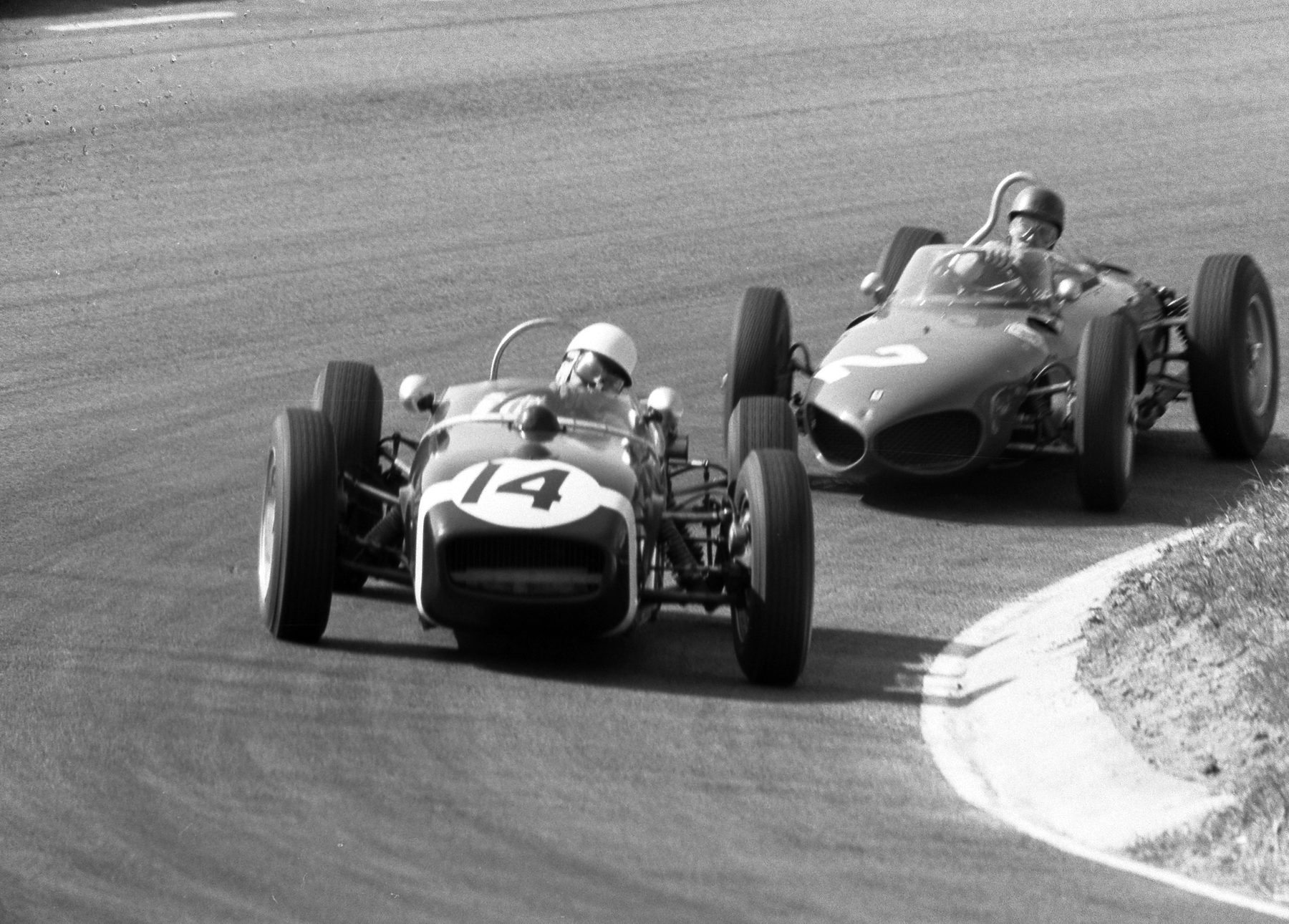 Two small open-wheeled racing cars take a bend close together: the leading one, nearer the camera, numbered 14, its driver in a white helmet; the other numbered 2, its driver in a dark helmet.