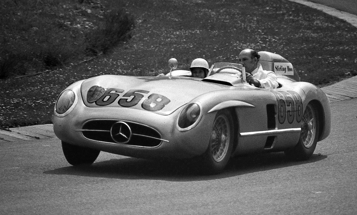A bare-headed man in a white shirt drives an open, streamlined sports-racing car numbered 658 along a track beside a grassy bank; a passenger in a pale helmet sits beside him.