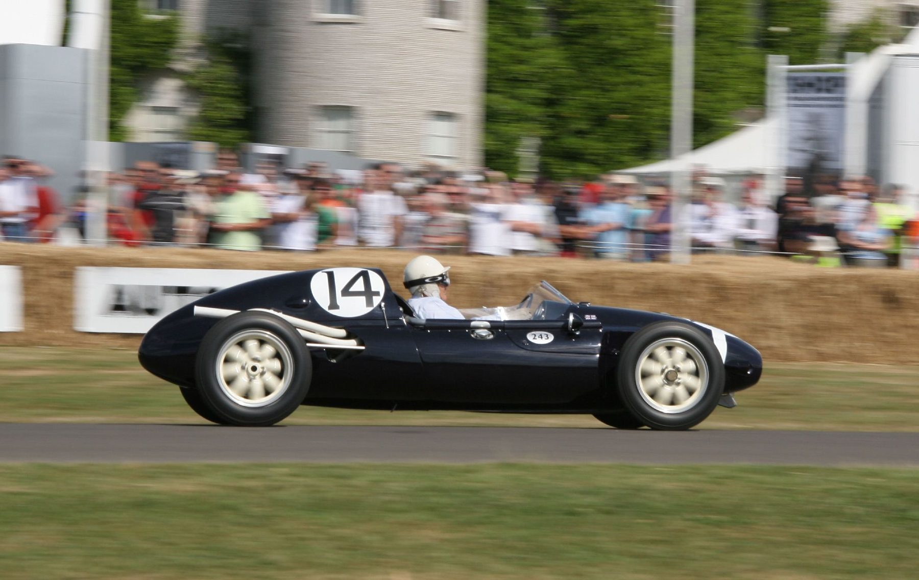 A driver in a white peaked helmet and goggles drives a small dark blue rear-engined racing car, numbered 14, past straw bales and a crowd of spectators.