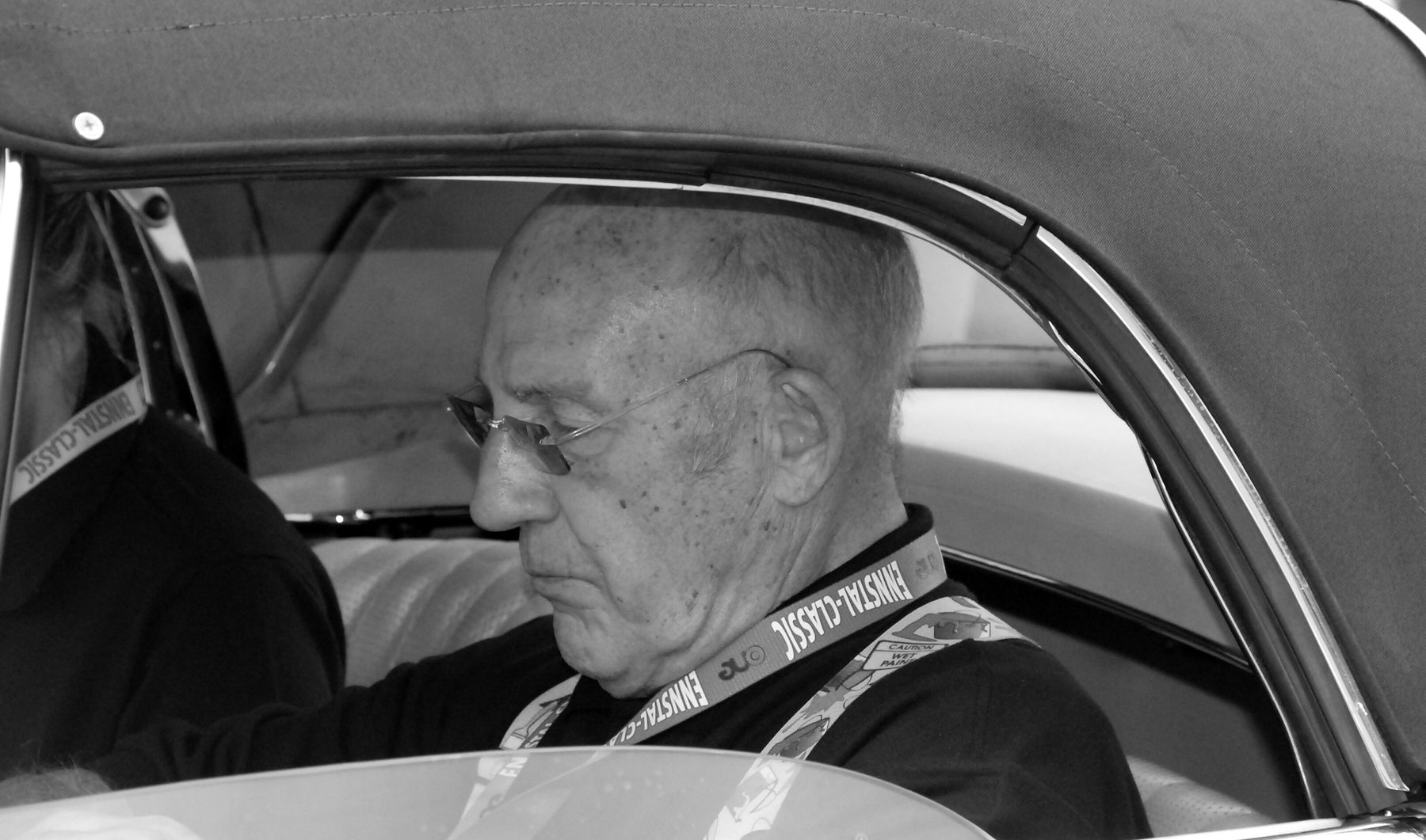 An elderly man in glasses and a dark top, a lanyard round his neck, sits in a car under its raised soft top and looks down; a second person sits partly hidden beside him at the left.